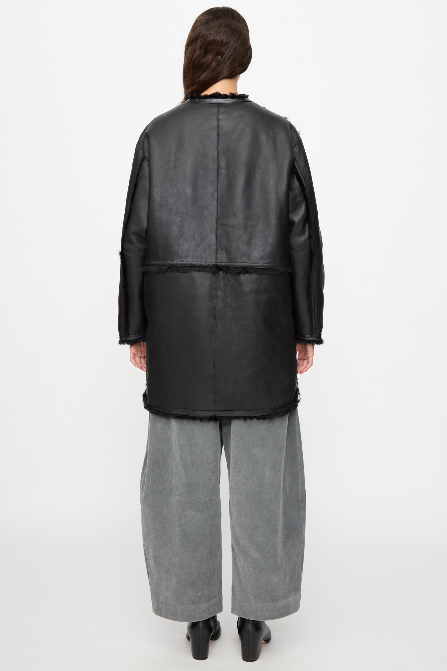A person stands with their back to the camera against a plain light background, wearing the Milena Silvano Sheepskin Reversible Liner in Black, styled with wide gray pants and black shoes.