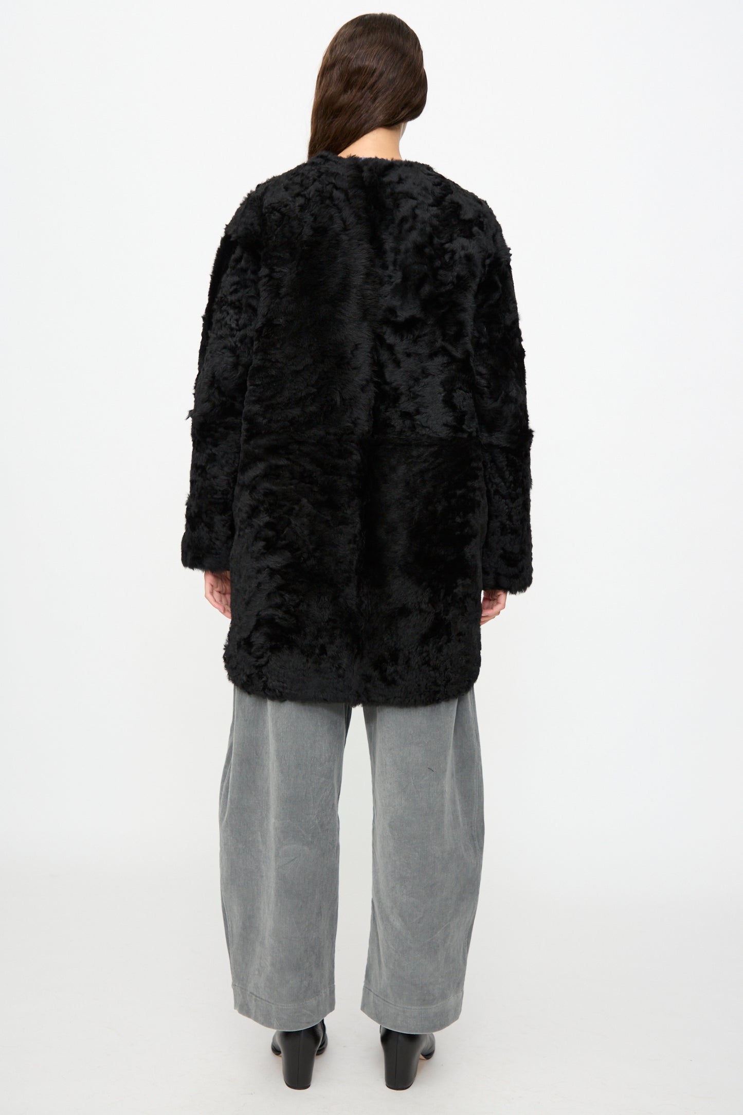 A person stands facing away, wearing the Milena Silvano Sheepskin Reversible Liner in Black, paired with wide gray pants and black heeled boots against a plain white background.