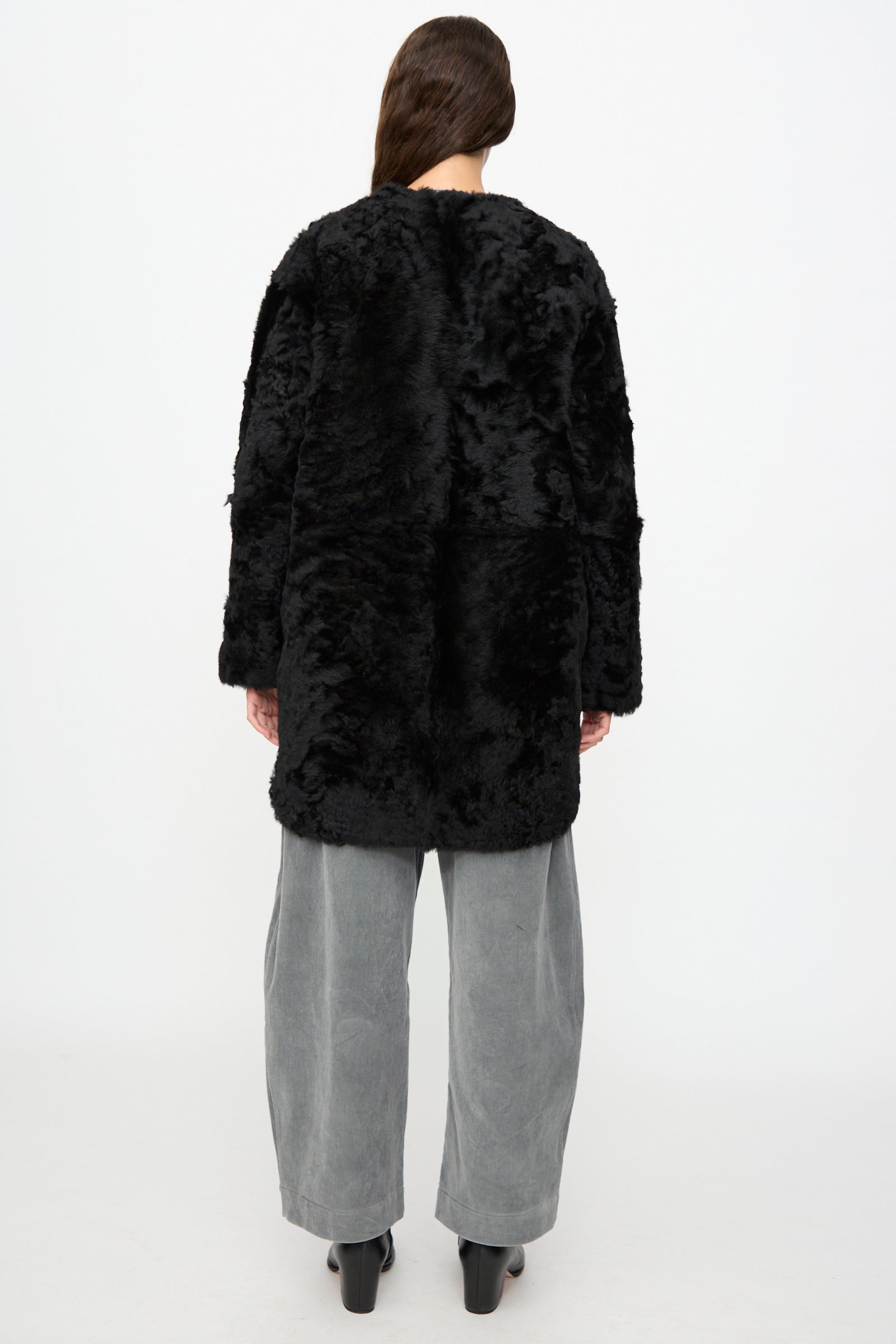A person stands facing away, wearing the Milena Silvano Sheepskin Reversible Liner in Black, paired with wide gray pants and black heeled boots against a plain white background.