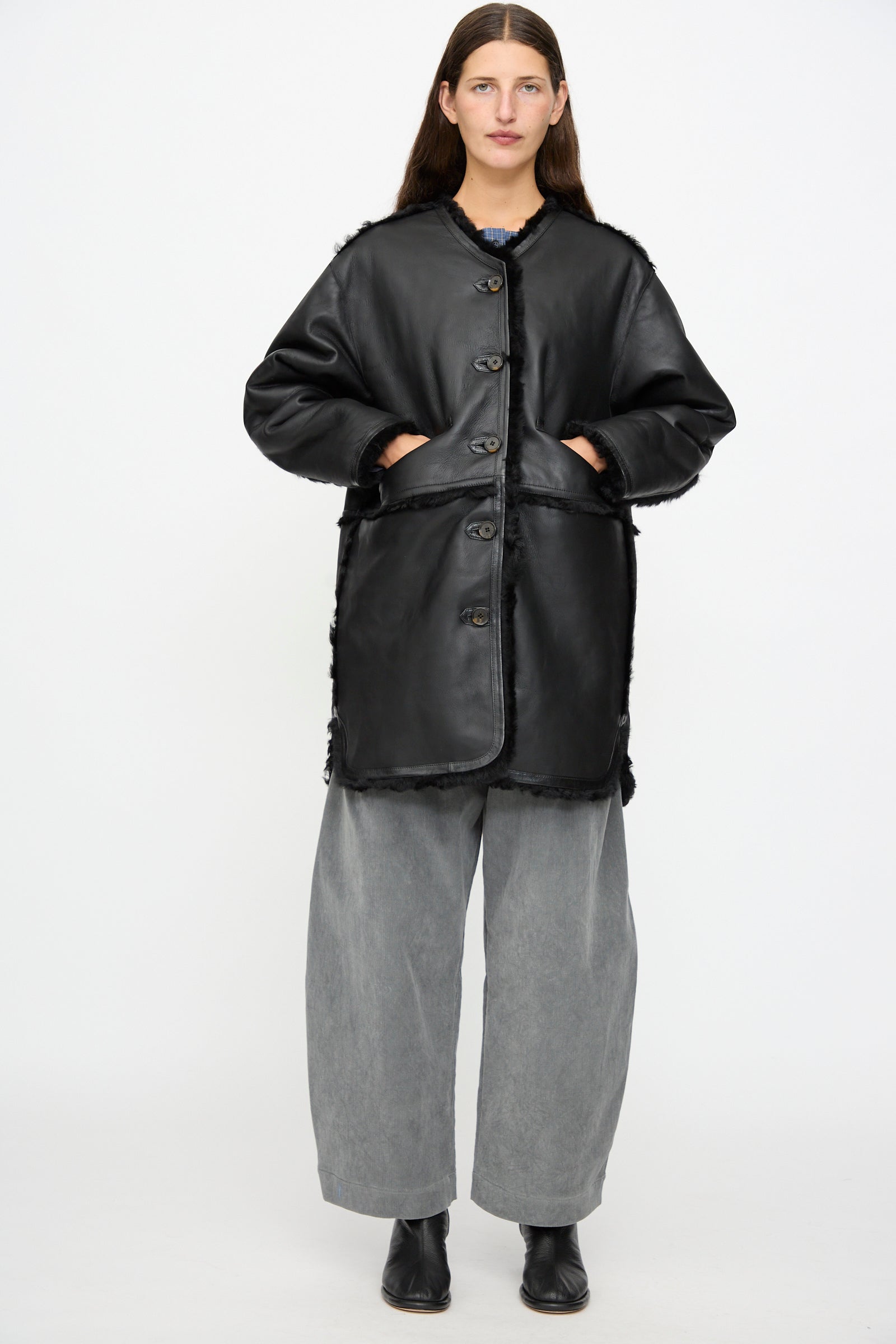 A person stands against a plain white background wearing the Milena Silvano Sheepskin Reversible Liner in Black, styled with hands in pockets, wide gray pants, and black boots.