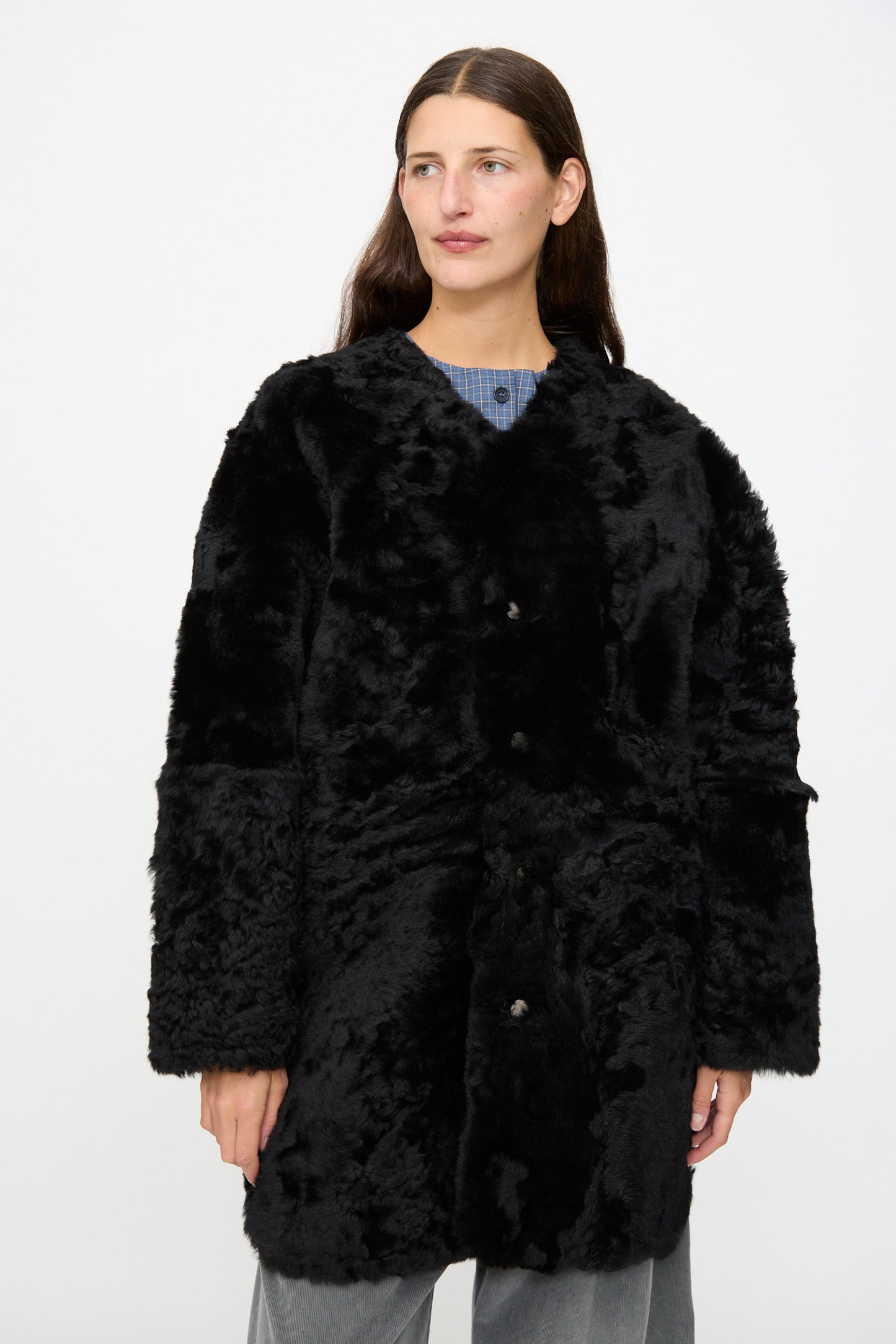 A person with long brown hair wears the Milena Silvano Sheepskin Reversible Liner in Black over a blue top and gray pants, standing against a plain white background.