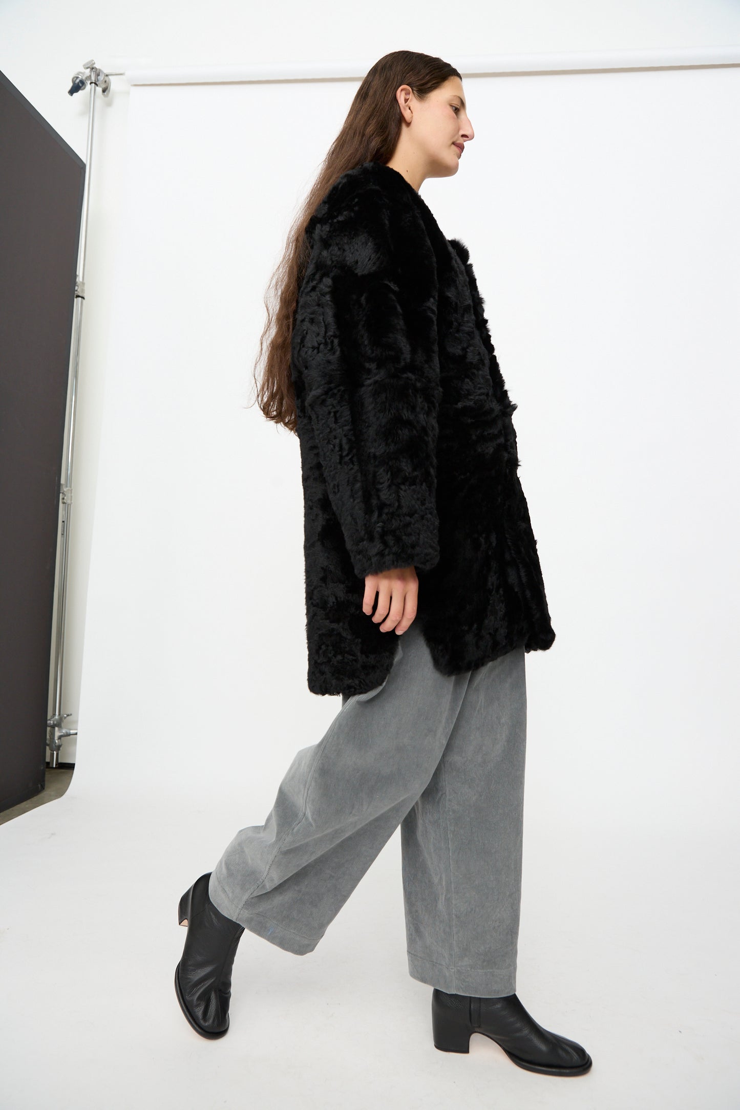 A person with long brown hair wears the Milena Silvano Sheepskin Reversible Liner in Black, gray wide-leg pants, and black ankle boots while walking in a white studio setting.