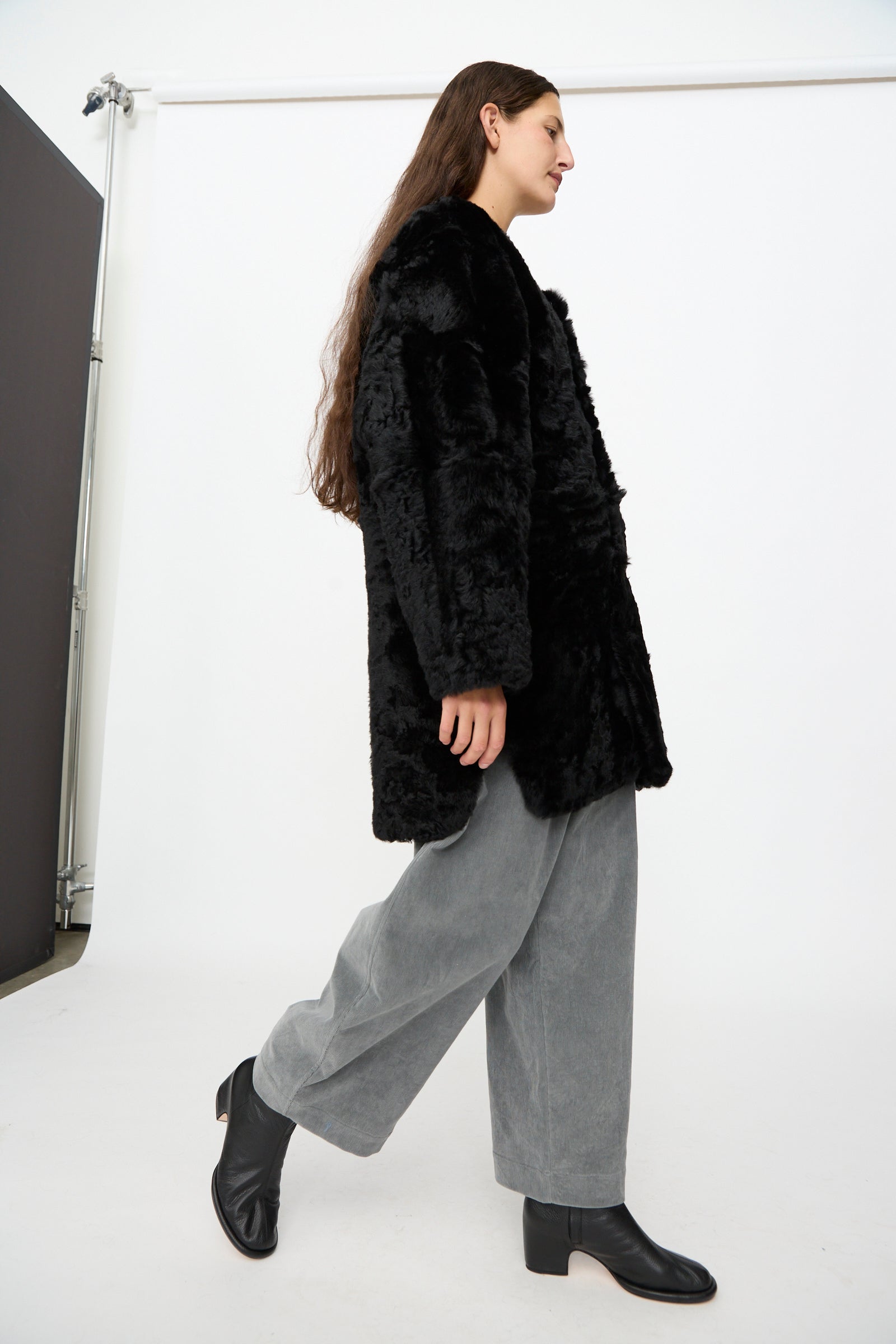 A person with long brown hair wears the Milena Silvano Sheepskin Reversible Liner in Black, gray wide-leg pants, and black ankle boots while walking in a white studio setting.
