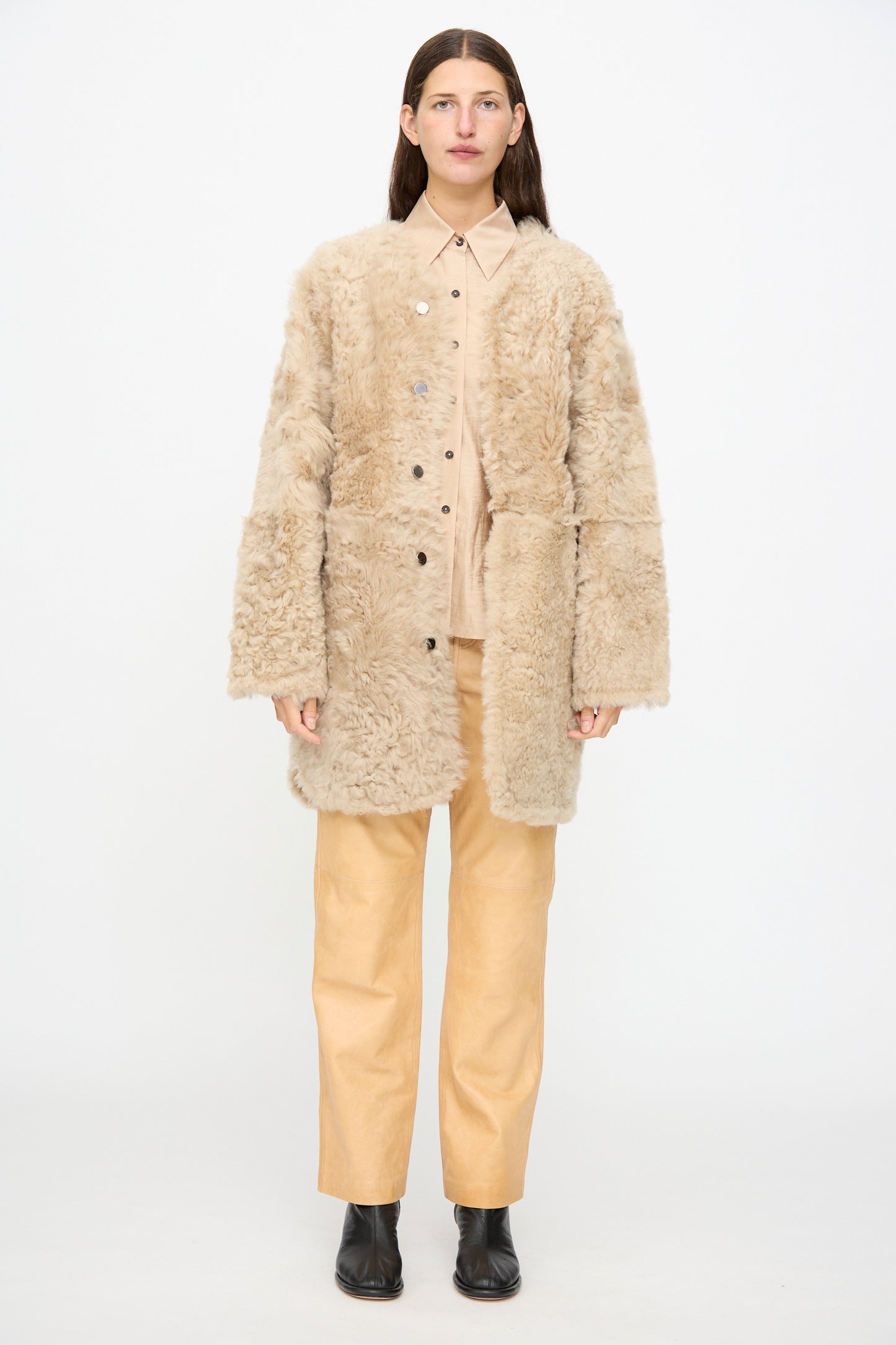 A person stands facing forward against a plain white background, wearing the Milena Silvano Sheepskin Reversible Liner in Oat with its beige exterior and black leather interior, paired with matching pants, a button-up shirt, and black ankle boots.