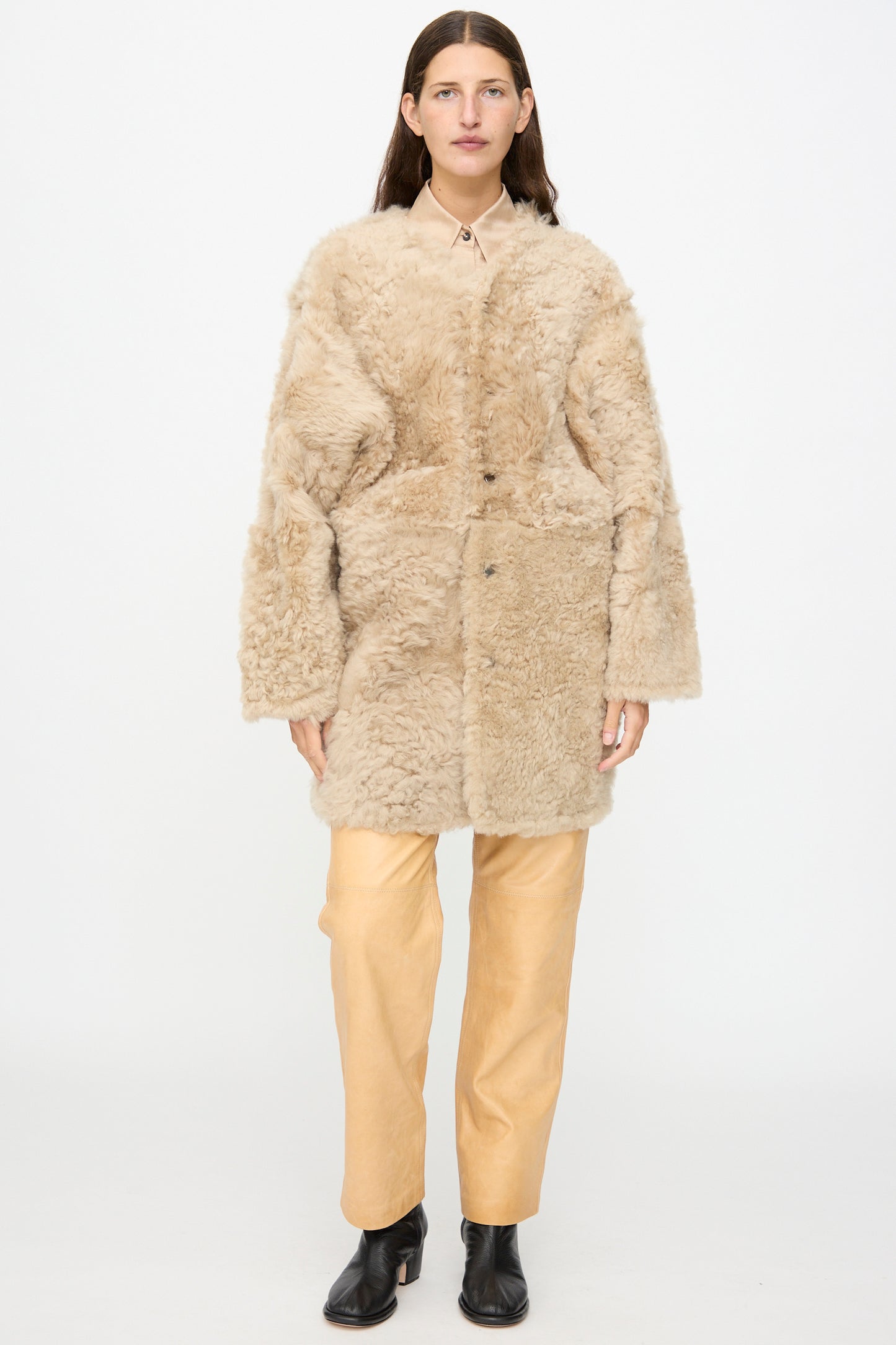 A person stands against a white background wearing the Milena Silvano Sheepskin Reversible Liner in Oat—a beige faux fur coat reminiscent of a reversible sheepskin jacket with curly sheepskin detailing—layered over a beige shirt and light tan pants, paired with black ankle boots.