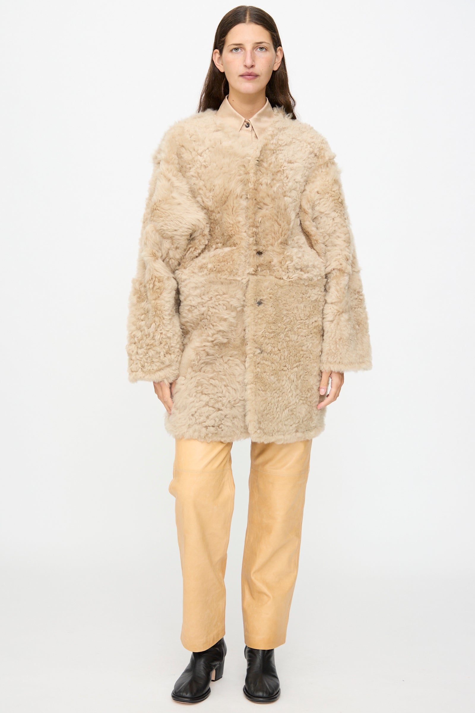 A person stands against a white background wearing the Milena Silvano Sheepskin Reversible Liner in Oat—a beige faux fur coat reminiscent of a reversible sheepskin jacket with curly sheepskin detailing—layered over a beige shirt and light tan pants, paired with black ankle boots.