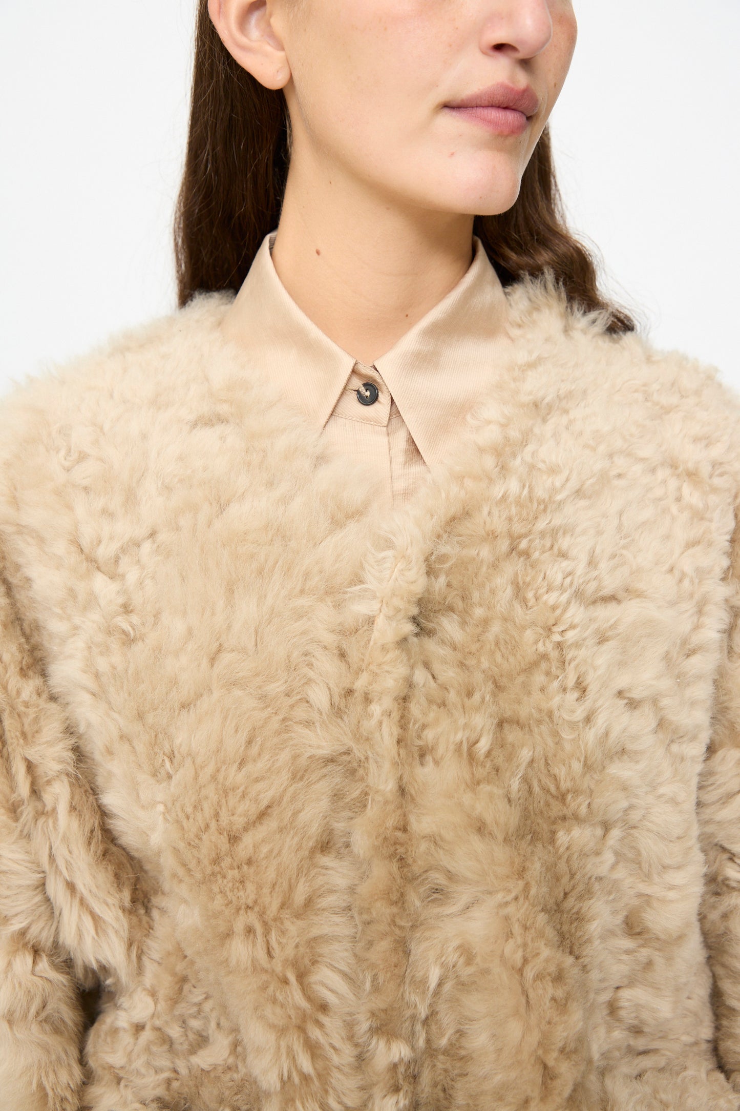 A person wearing the Milena Silvano Sheepskin Reversible Liner in Oat styled as a jacket over a buttoned-up light tan collared shirt, photographed from shoulders to nose.