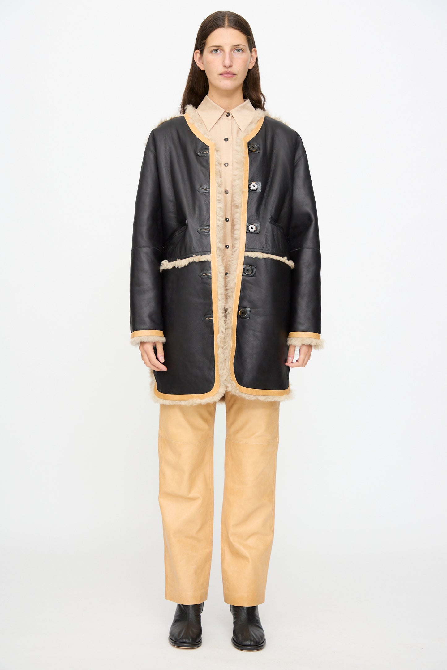 A person stands against a white background, wearing the Milena Silvano Sheepskin Reversible Liner in Oat—with a curly sheepskin exterior and black leather interior—styled over a beige button-up shirt and beige pants, paired with black ankle boots.