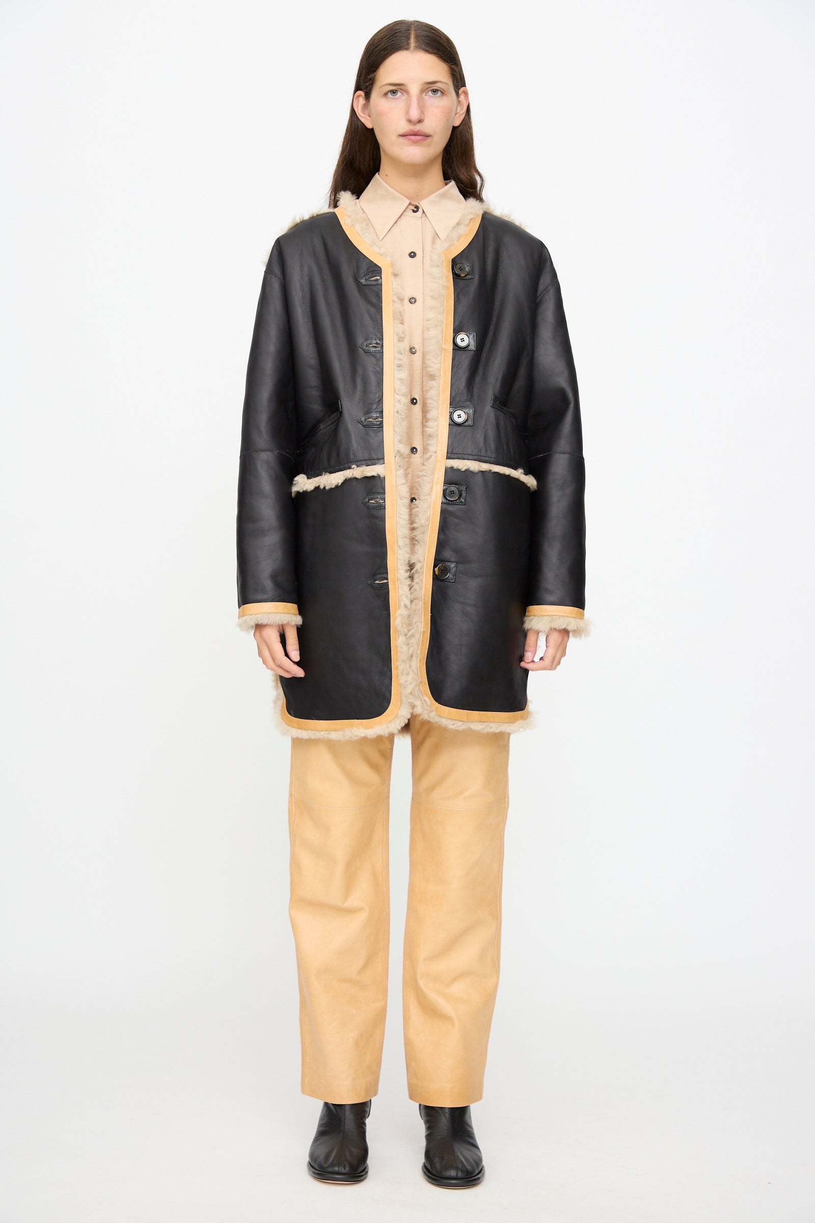 A person stands against a white background, wearing the Milena Silvano Sheepskin Reversible Liner in Oat—with a curly sheepskin exterior and black leather interior—styled over a beige button-up shirt and beige pants, paired with black ankle boots.