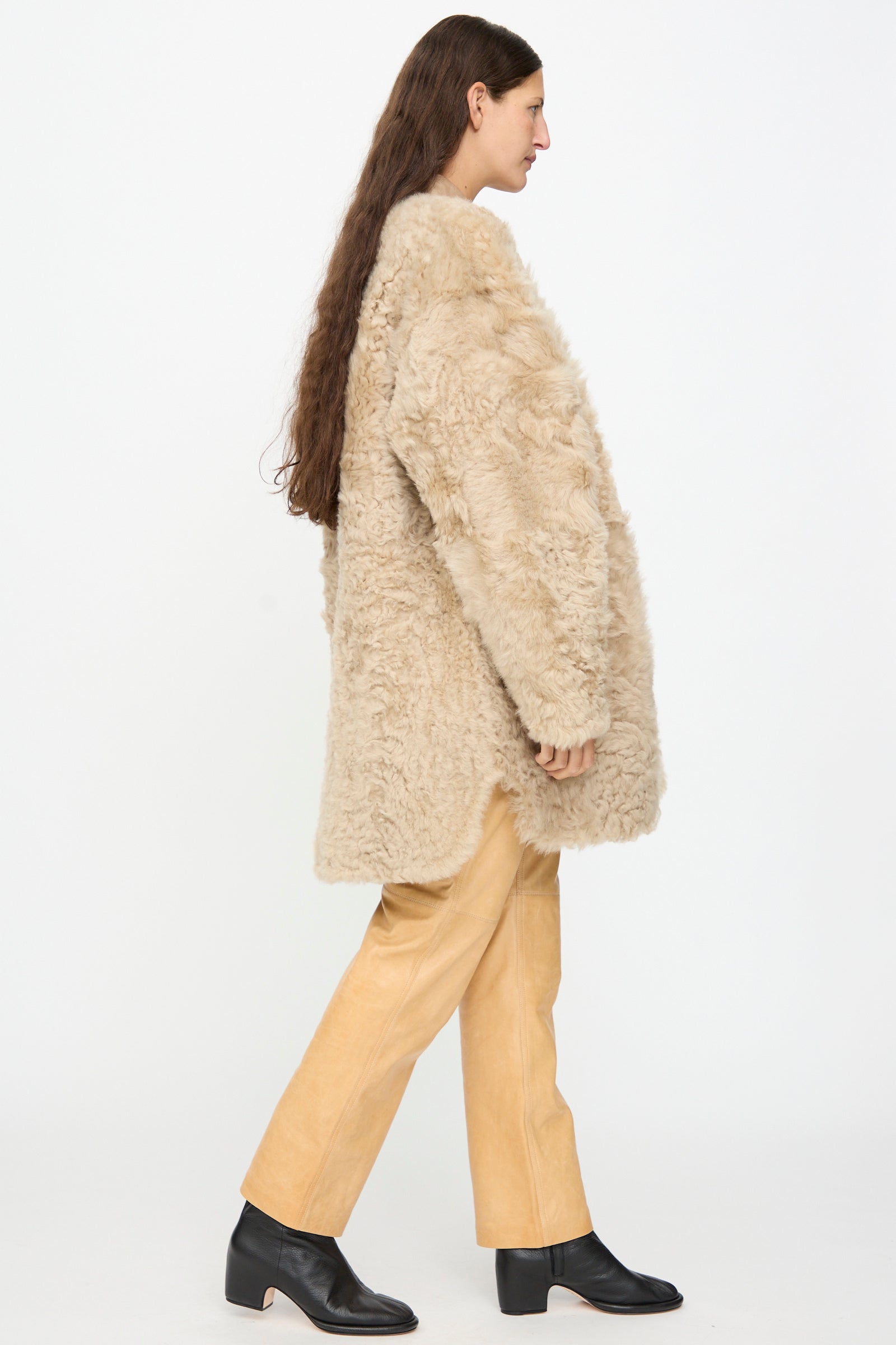 A person in profile wears the Milena Silvano Sheepskin Reversible Liner in Oat, featuring a beige exterior and black leather interior, paired with tan pants and black ankle boots, standing against a plain white background.