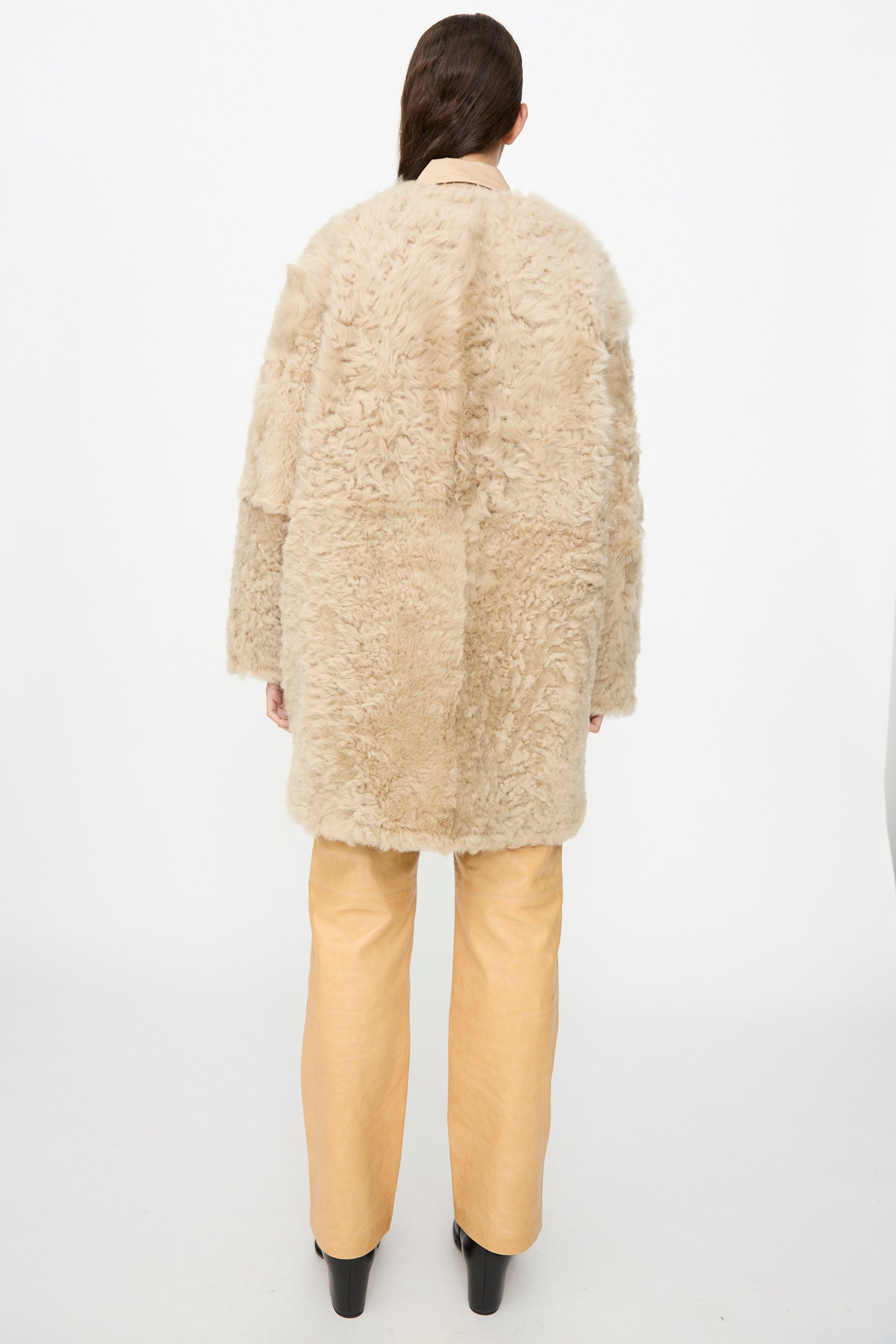 A person stands with their back to the camera, wearing the Milena Silvano Sheepskin Reversible Liner in Oat with curly sheepskin detailing, paired with tan wide-leg pants and black platform shoes, set against a plain white background.