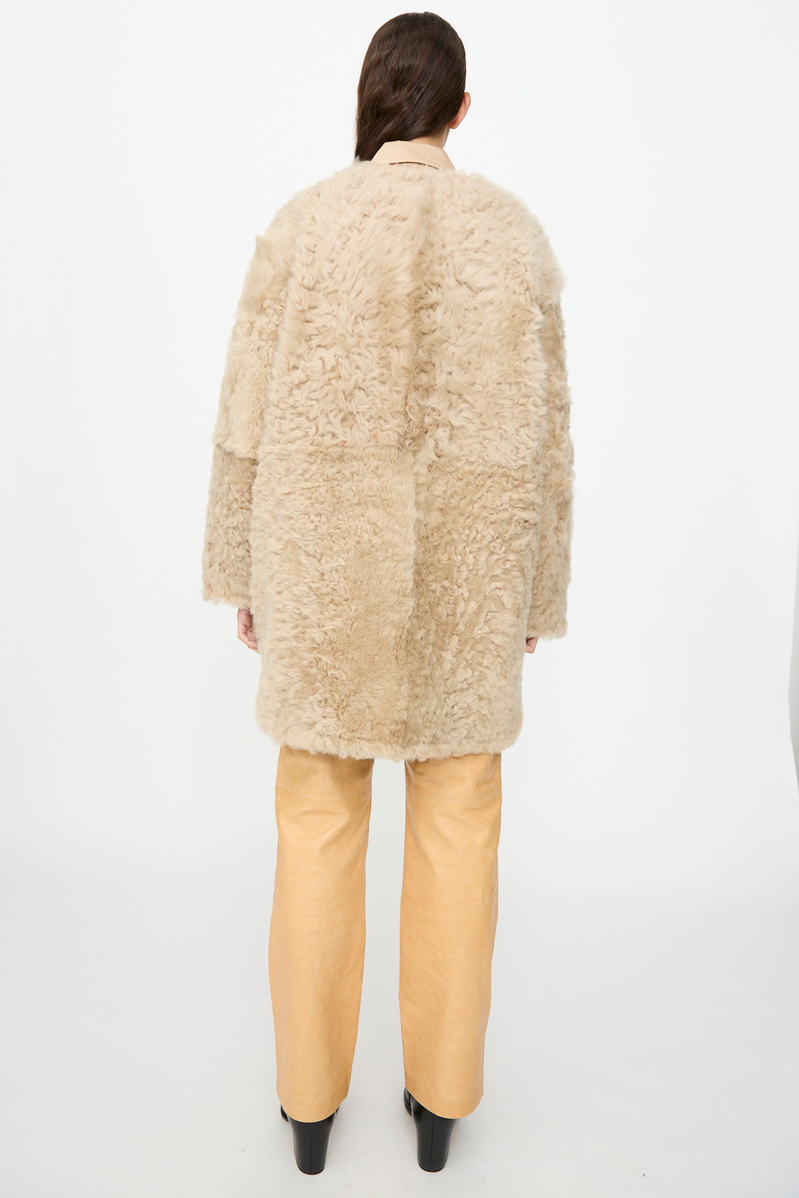 A person stands with their back to the camera, wearing the Milena Silvano Sheepskin Reversible Liner in Oat with curly sheepskin detailing, paired with tan wide-leg pants and black platform shoes, set against a plain white background.