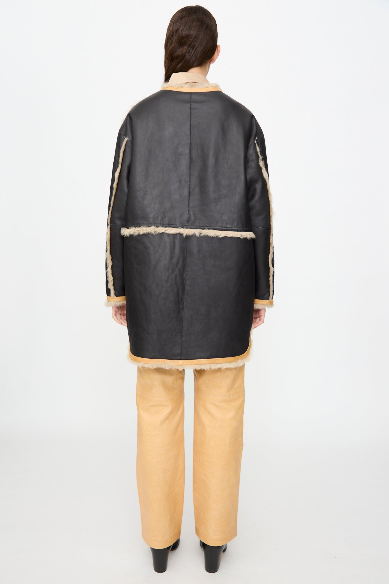 A person stands facing away, wearing the Milena Silvano Sheepskin Reversible Liner in Oat—featuring contrasting black and beige panels—paired with tan pants and black shoes against a plain white background.
