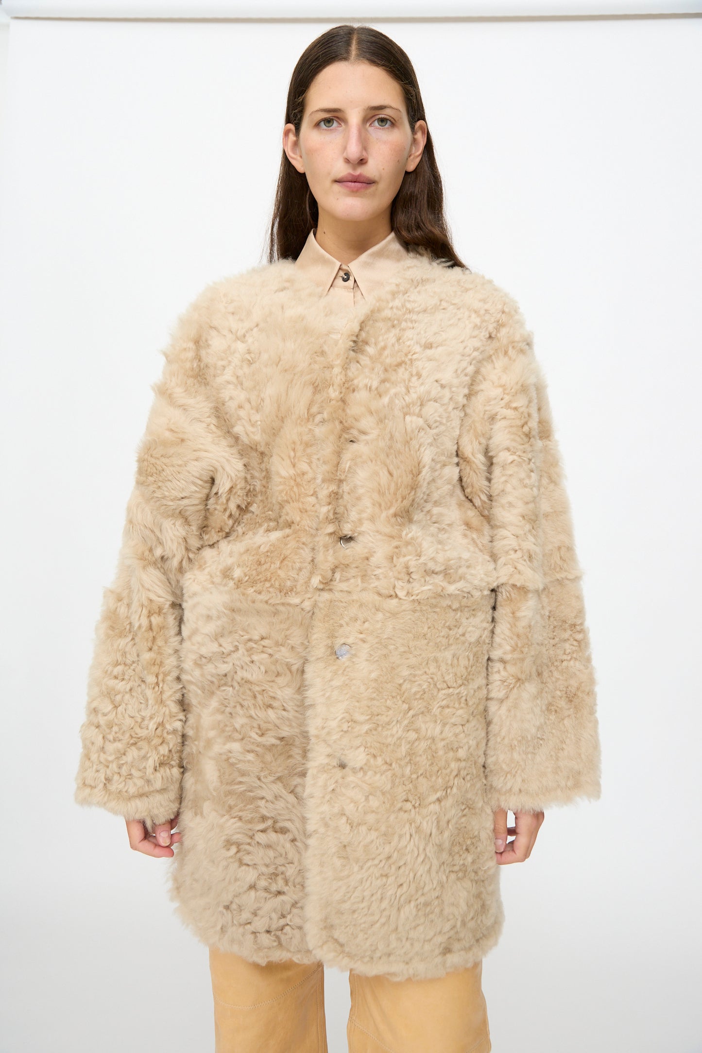 A woman wearing the Milena Silvano Sheepskin Reversible Liner in Oat over a collared shirt and tan pants stands against a plain white background.