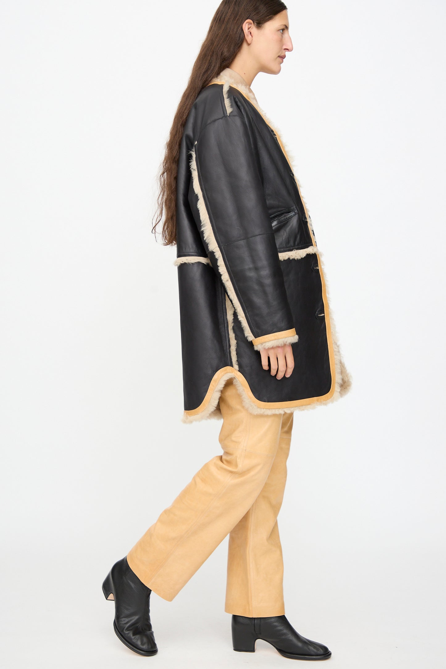 A person with long brown hair, dressed in the Milena Silvano Sheepskin Reversible Liner in Oat, beige pants, and black heeled boots, walks to the left against a plain white background.