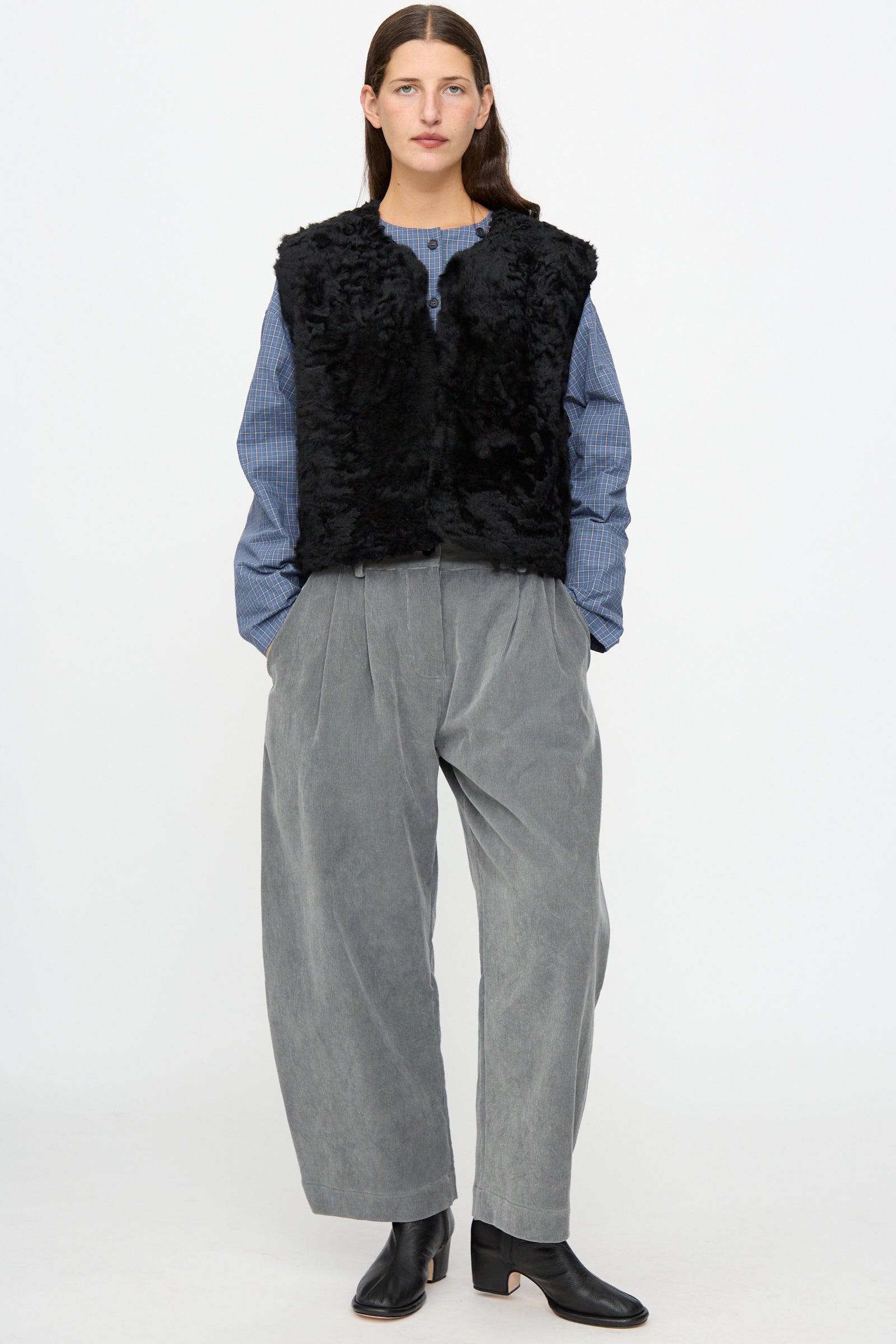 A person wearing the Milena Silvano Sheepskin Transition Vest in Black, paired with a blue checkered blouse, gray wide-leg pants, and black ankle boots, standing against a plain white background.