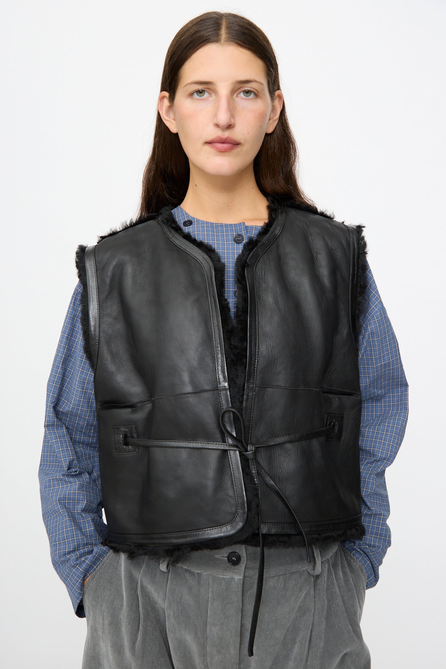 A woman wears the Milena Silvano Sheepskin Transition Vest in Black over a blue checkered shirt and gray trousers, standing against a plain light background.