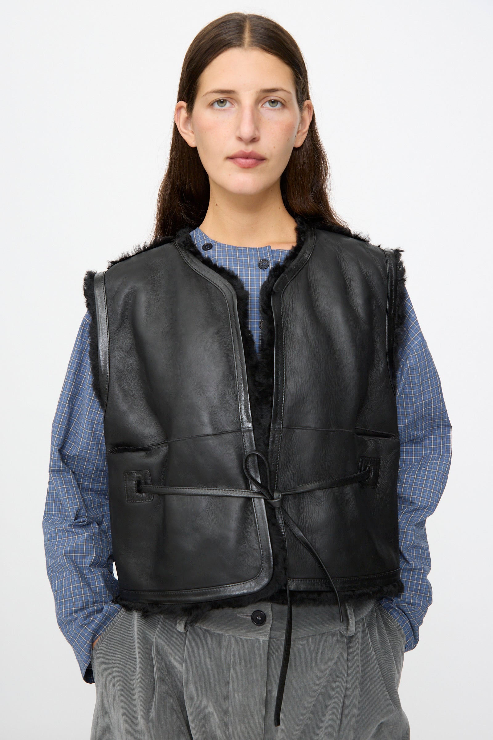 A woman wears the Milena Silvano Sheepskin Transition Vest in Black over a blue checkered shirt and gray trousers, standing against a plain light background.