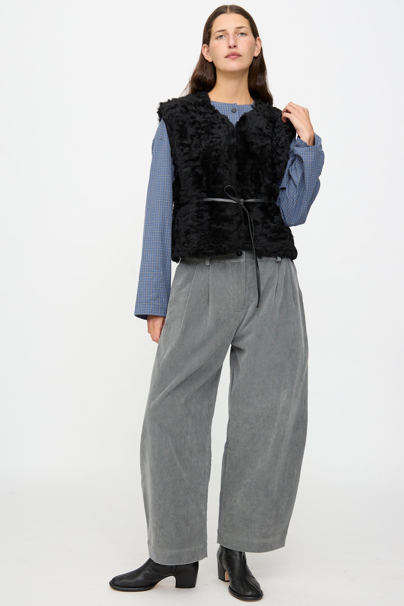 A person stands against a plain background wearing the Milena Silvano Sheepskin Transition Vest in Black, paired with a blue long-sleeve shirt, wide gray pants, and black ankle boots.