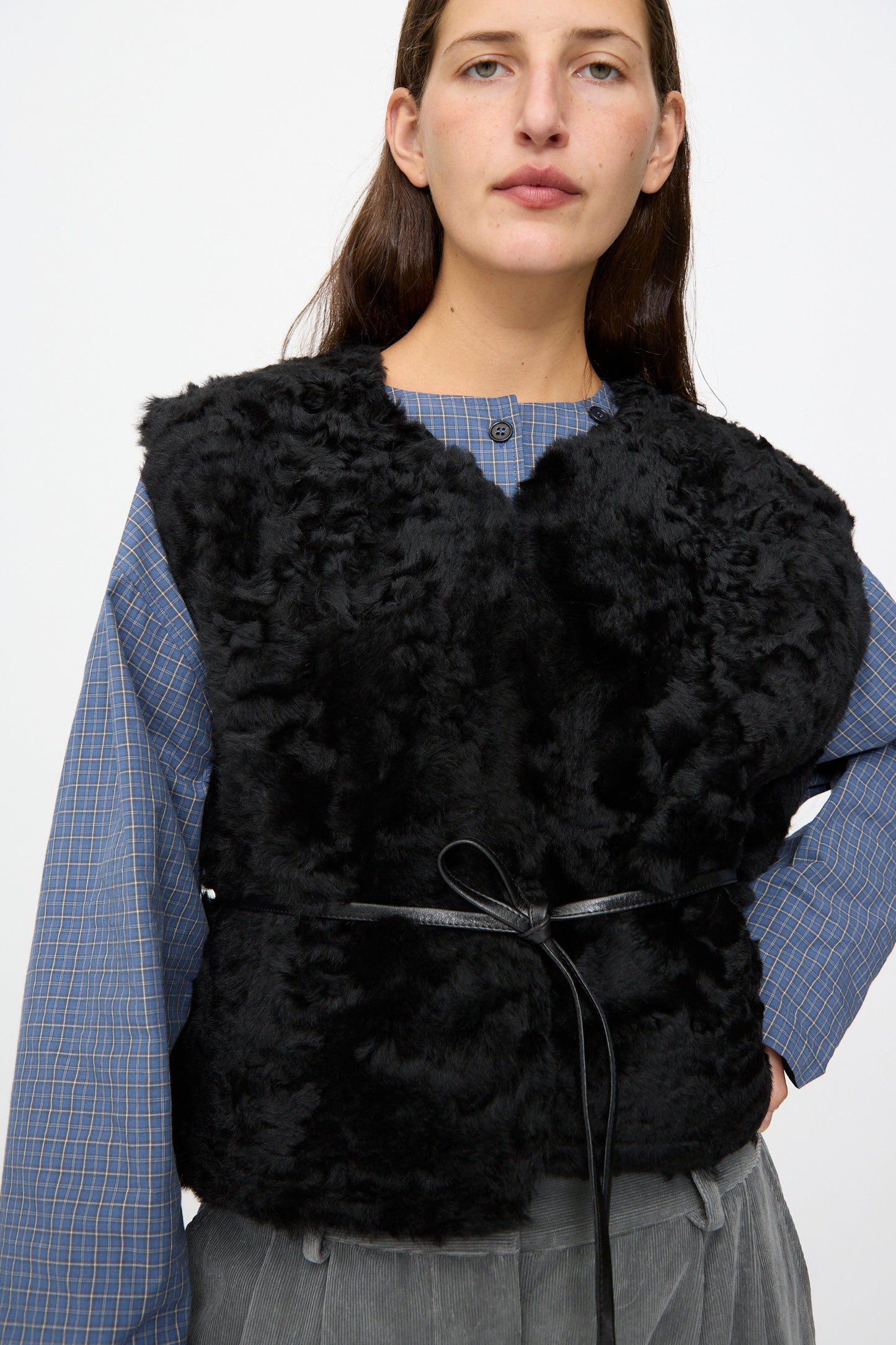 A person wears a blue plaid shirt, the Milena Silvano Sheepskin Transition Vest in Black, which features a reversible design and thin belt, paired with gray pants, standing against a plain light background.