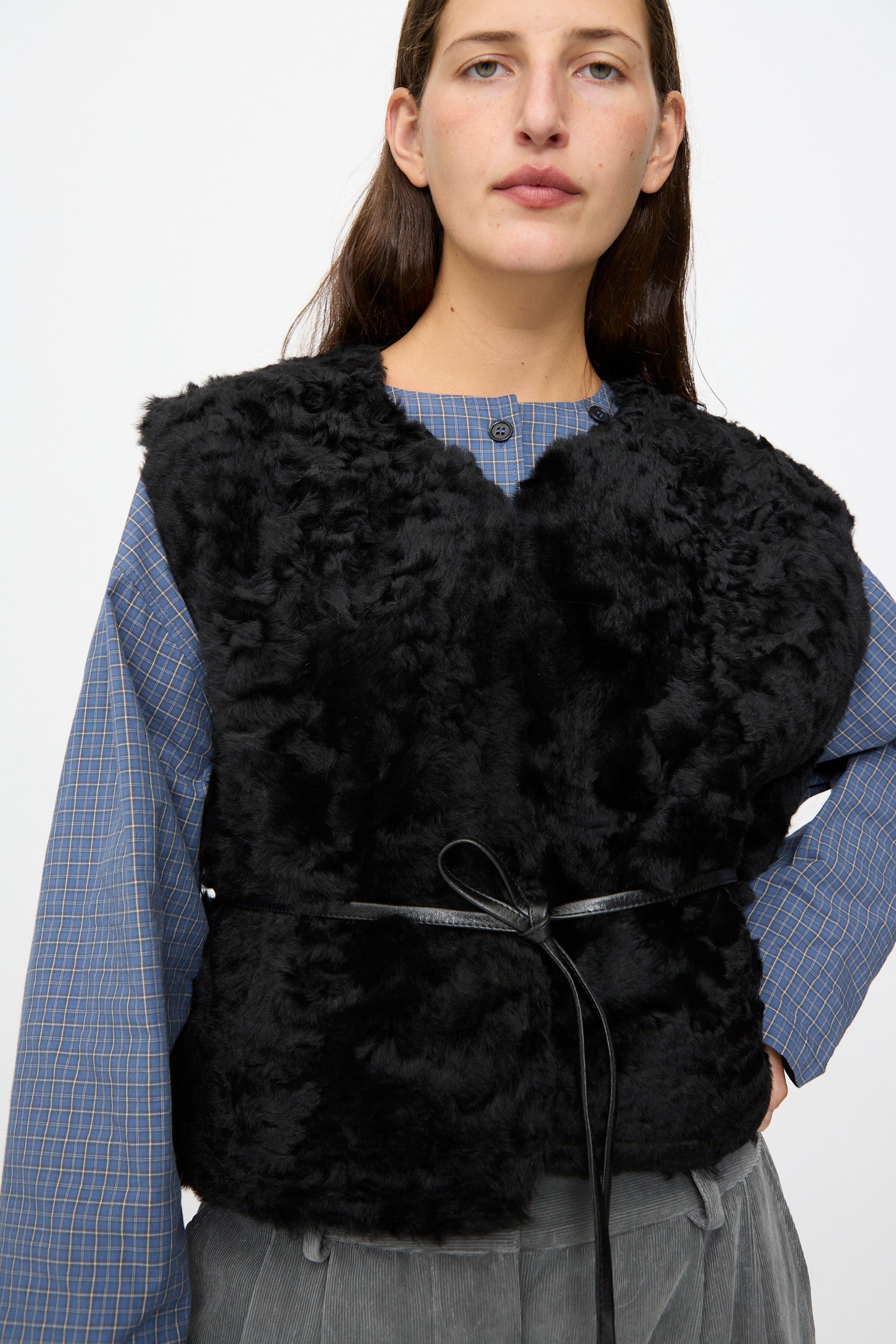 A person wears a blue plaid shirt, the Milena Silvano Sheepskin Transition Vest in Black, which features a reversible design and thin belt, paired with gray pants, standing against a plain light background.