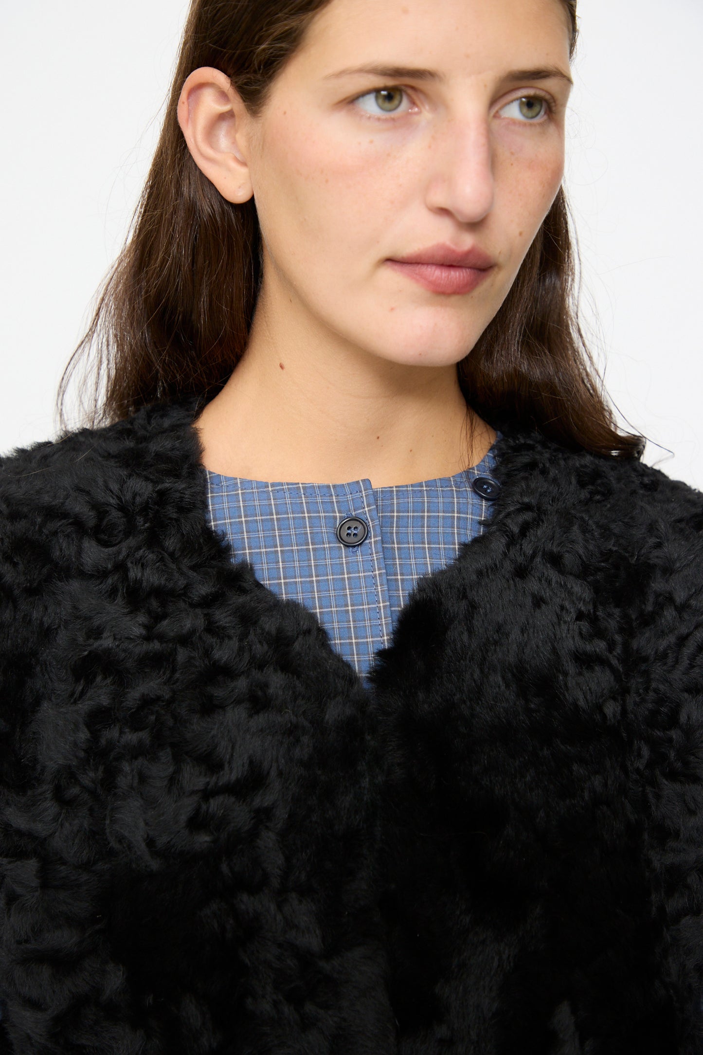 A person with long brown hair wears a black textured coat over a blue plaid shirt and the Milena Silvano Sheepskin Transition Vest in Black, facing slightly to the right against a plain background.