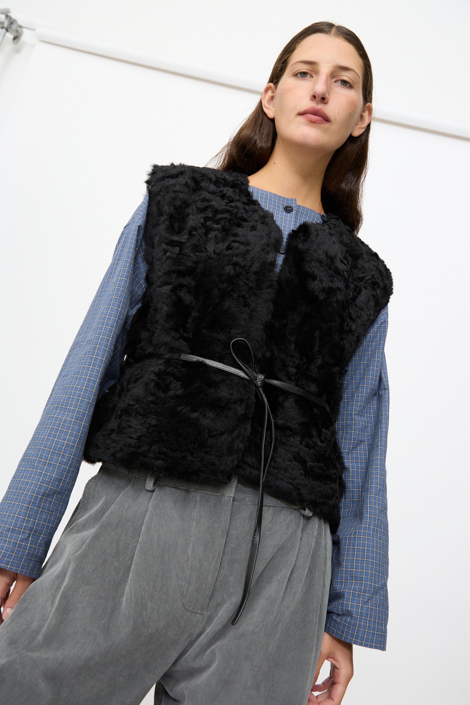A person wearing a blue checkered shirt, grey trousers, and the Milena Silvano Sheepskin Transition Vest in Black stands in front of a white background.