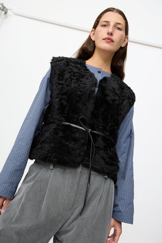 A person wearing a blue checkered shirt, grey trousers, and the Milena Silvano Sheepskin Transition Vest in Black stands in front of a white background.