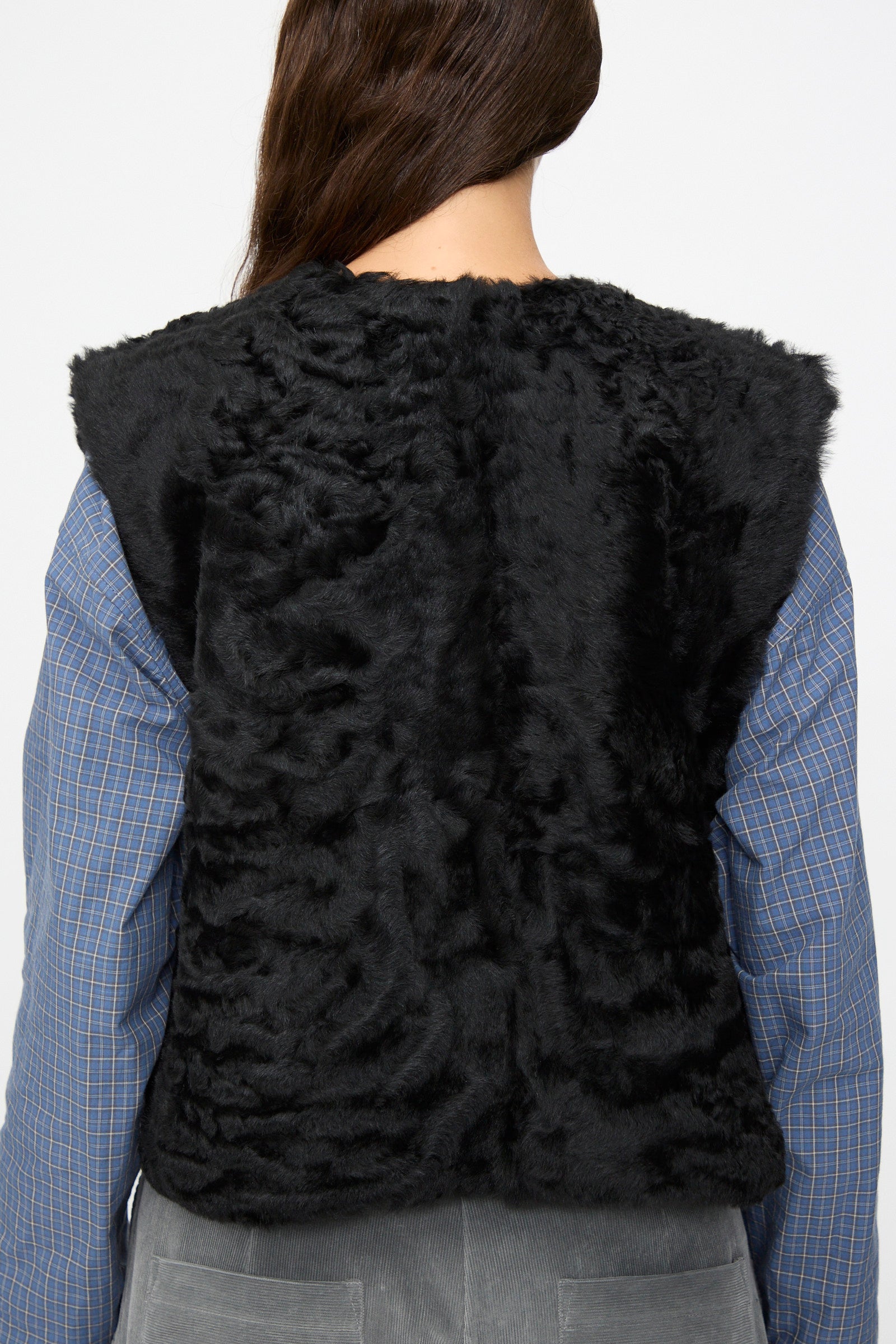 A person seen from behind wearing the Milena Silvano Sheepskin Transition Vest in Black over a blue plaid shirt and gray pants.