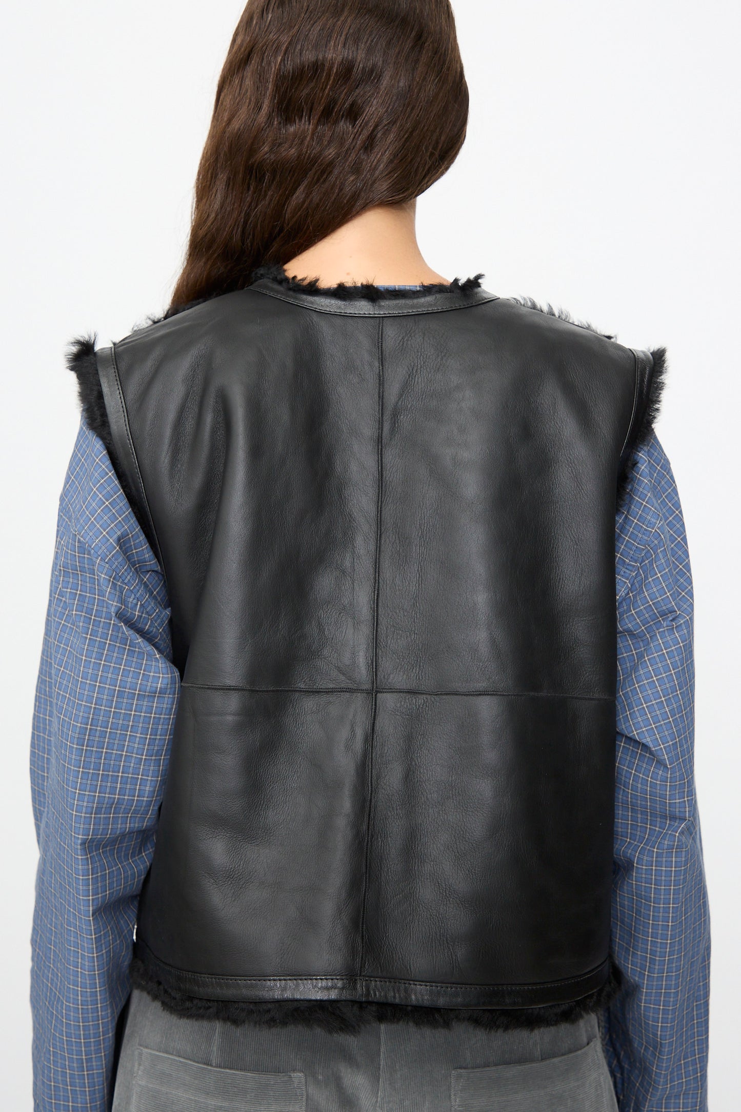 A person with long brown hair is shown from behind, wearing a blue plaid shirt layered with the Sheepskin Transition Vest in Black by Milena Silvano. The sleek black vest by Milena Silvano adds an edgy touch to the casual outfit.