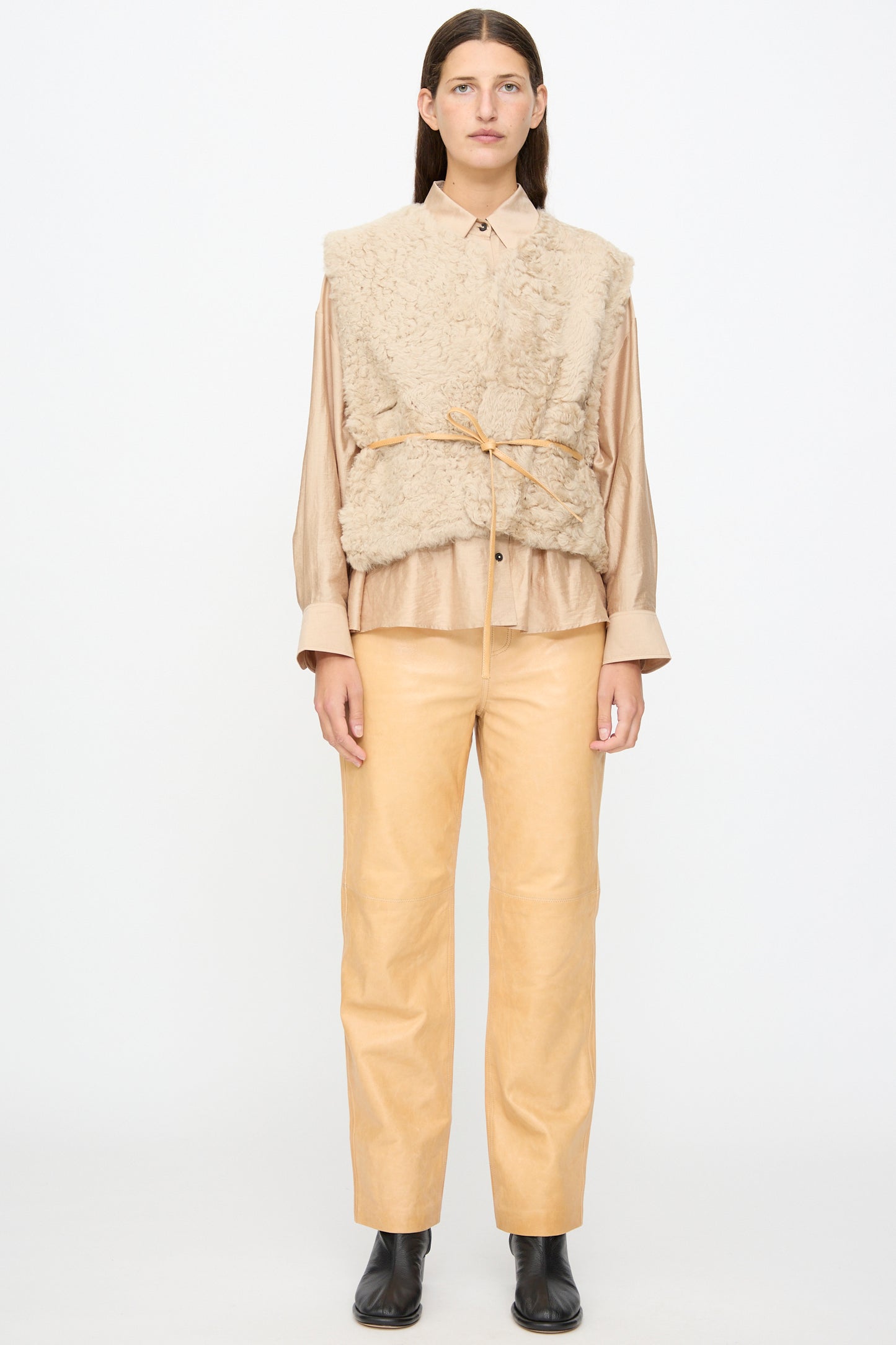 A person wears the Milena Silvano Sheepskin Transition Vest in Oat over a collared shirt, paired with tan pants and black boots, standing against a plain white background.