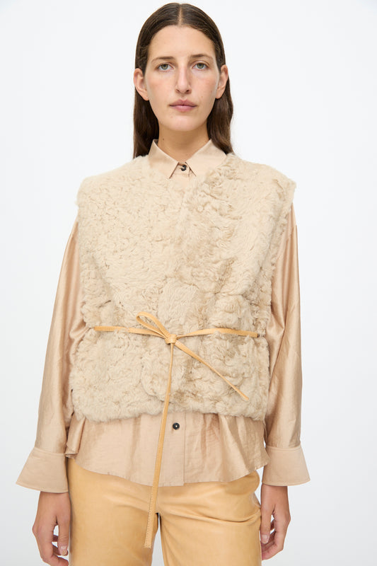 A woman wears the Milena Silvano Sheepskin Transition Vest in Oat over a light tan button-up shirt and matching relaxed-fit pants, tied at the waist with a thin belt, standing against a plain white background.