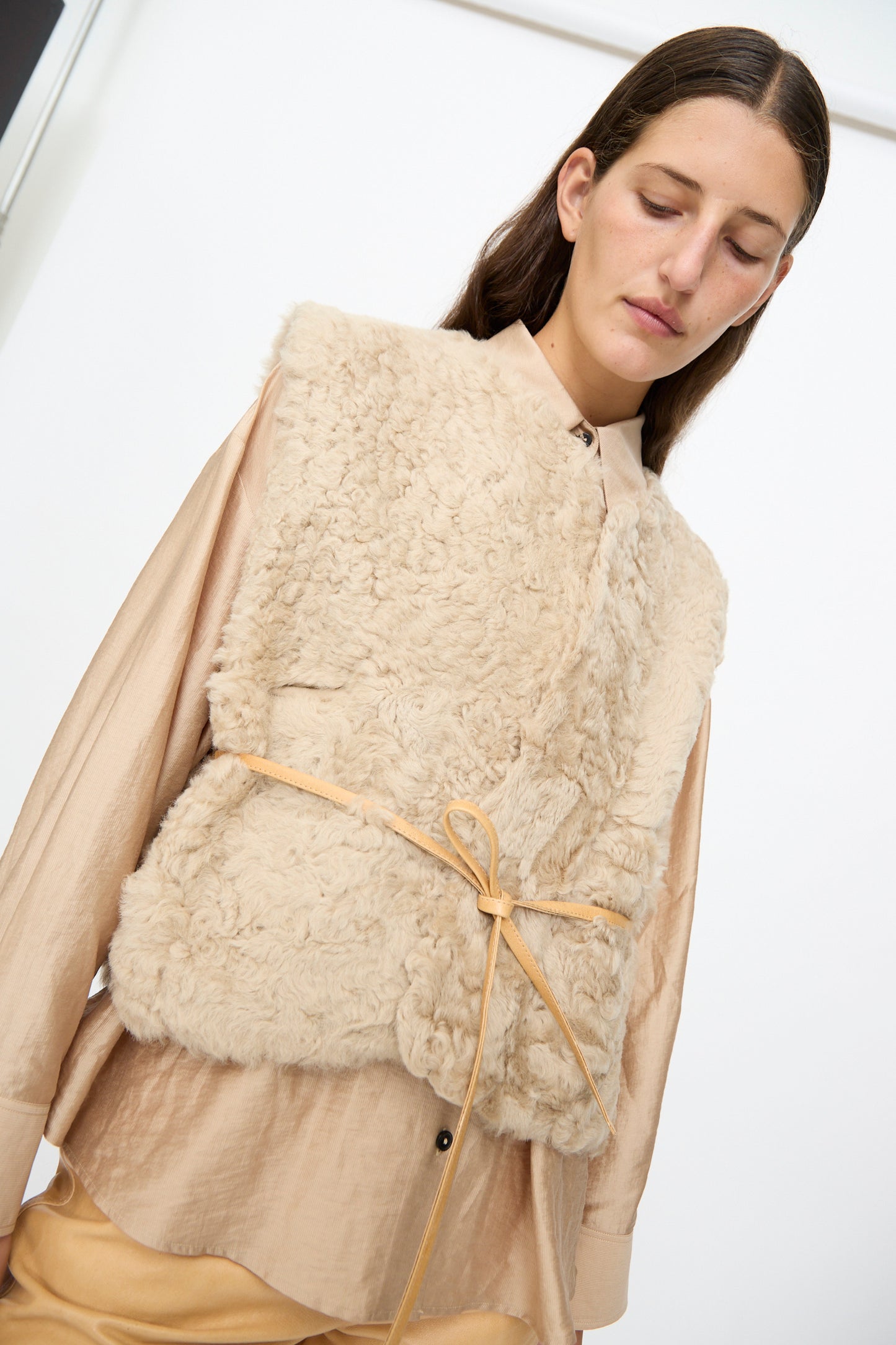 A person wearing the Milena Silvano Sheepskin Transition Vest in Oat over a light tan shirt, cinched at the waist with a thin belt, stands against a plain white background.