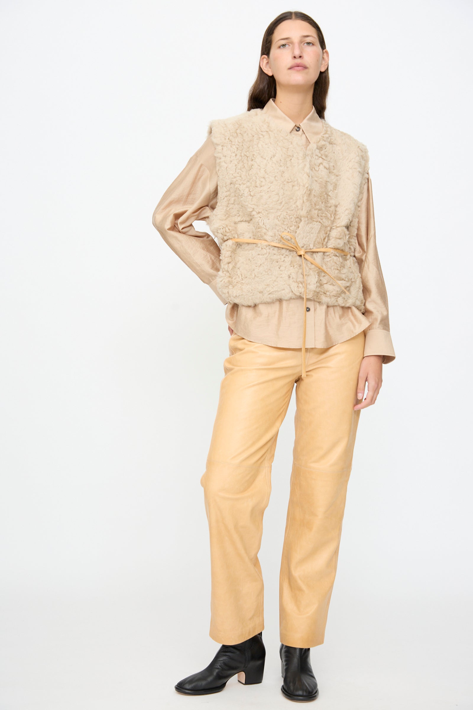 A person stands against a plain background, wearing a beige shirt, tan pants, black ankle boots, and a thin tan belt tied around the waist. Layered on top is the Milena Silvano Sheepskin Transition Vest in Oat.
