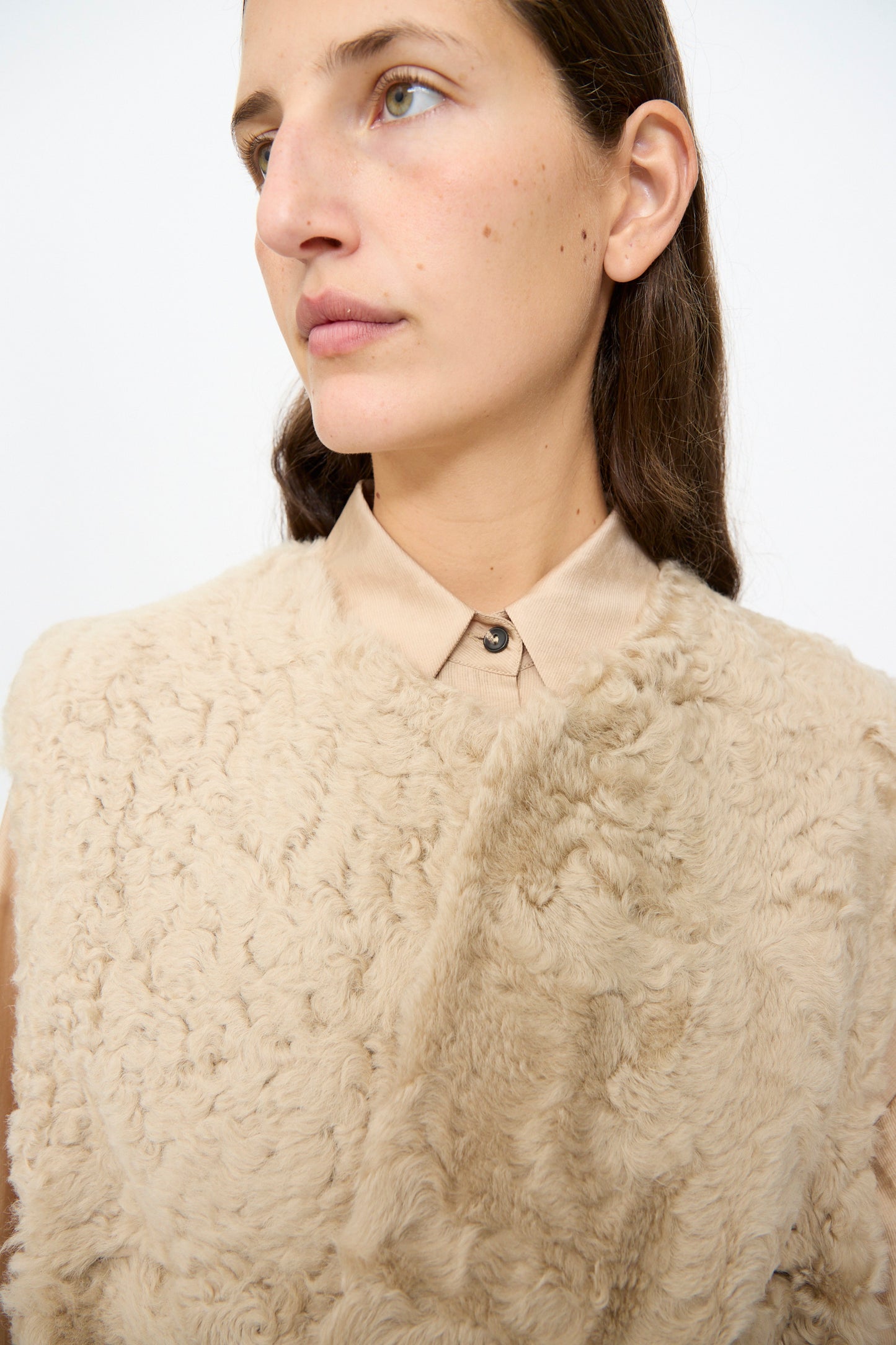 A person with long brown hair wears the Milena Silvano Sheepskin Transition Vest in Oat over a tan collared shirt, looking slightly to the side against a plain, light background.