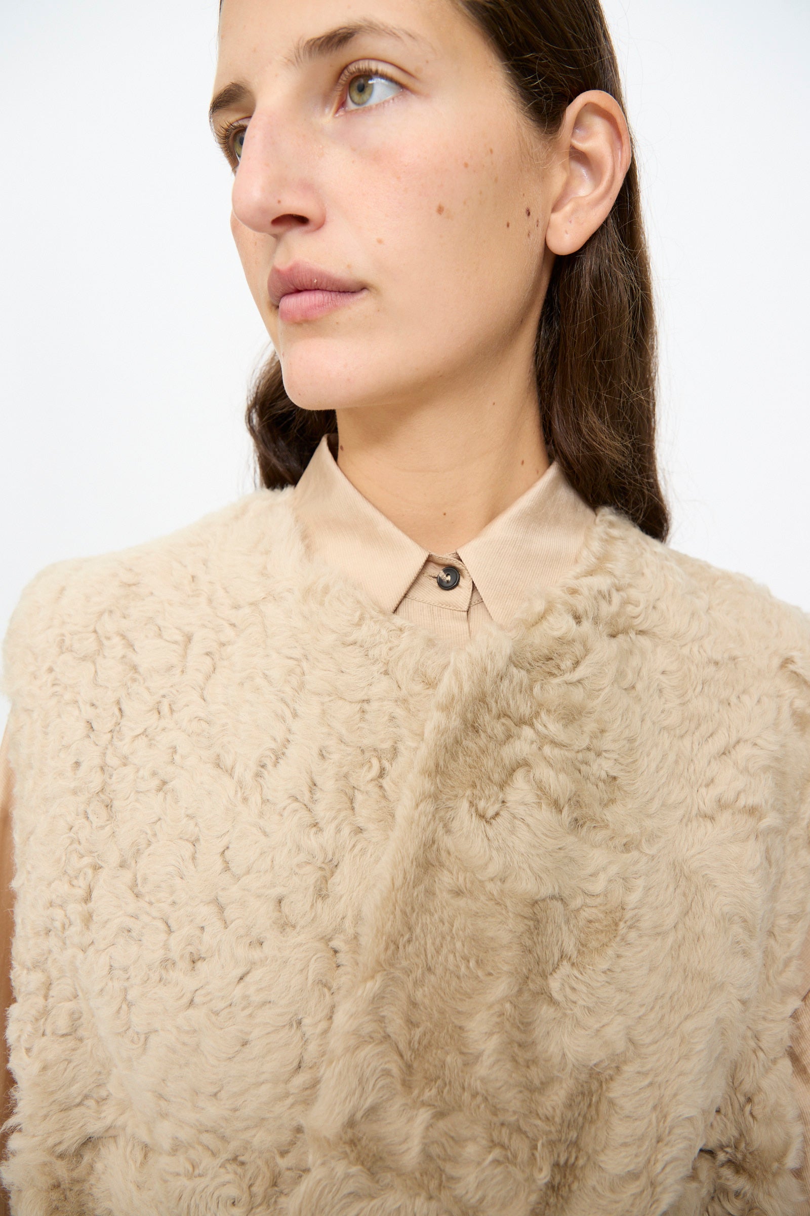 A person with long brown hair wears the Milena Silvano Sheepskin Transition Vest in Oat over a tan collared shirt, looking slightly to the side against a plain, light background.
