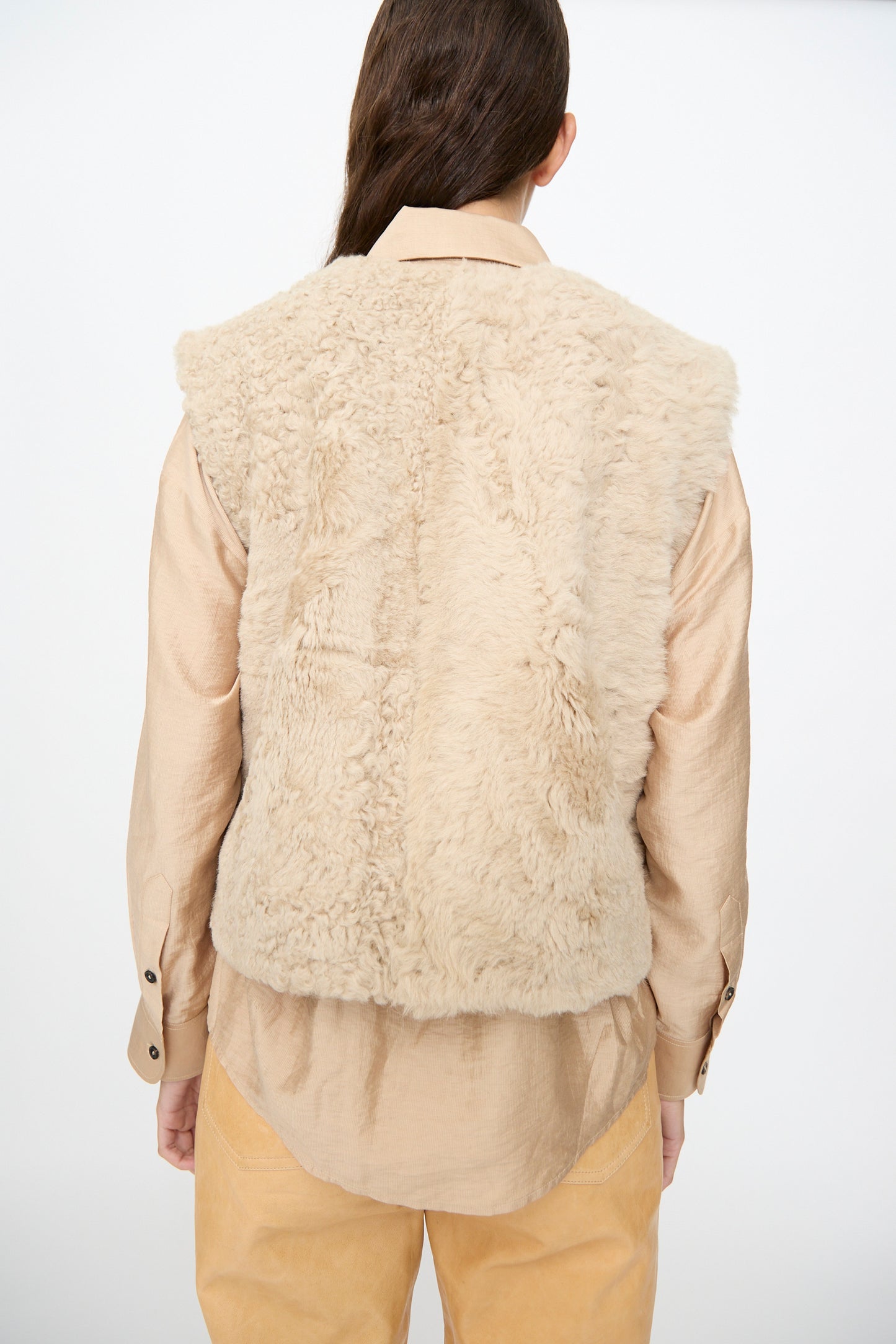 A person with long brown hair faces away, wearing the Milena Silvano Sheepskin Transition Vest in Oat—a relaxed-fit beige sheepskin vest—layered over a light tan shirt and tan pants, standing against a plain white background.