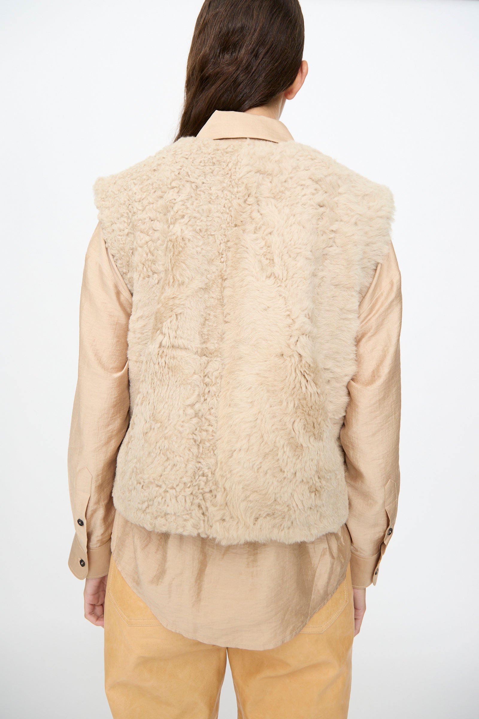 A person with long brown hair faces away, wearing the Milena Silvano Sheepskin Transition Vest in Oat—a relaxed-fit beige sheepskin vest—layered over a light tan shirt and tan pants, standing against a plain white background.