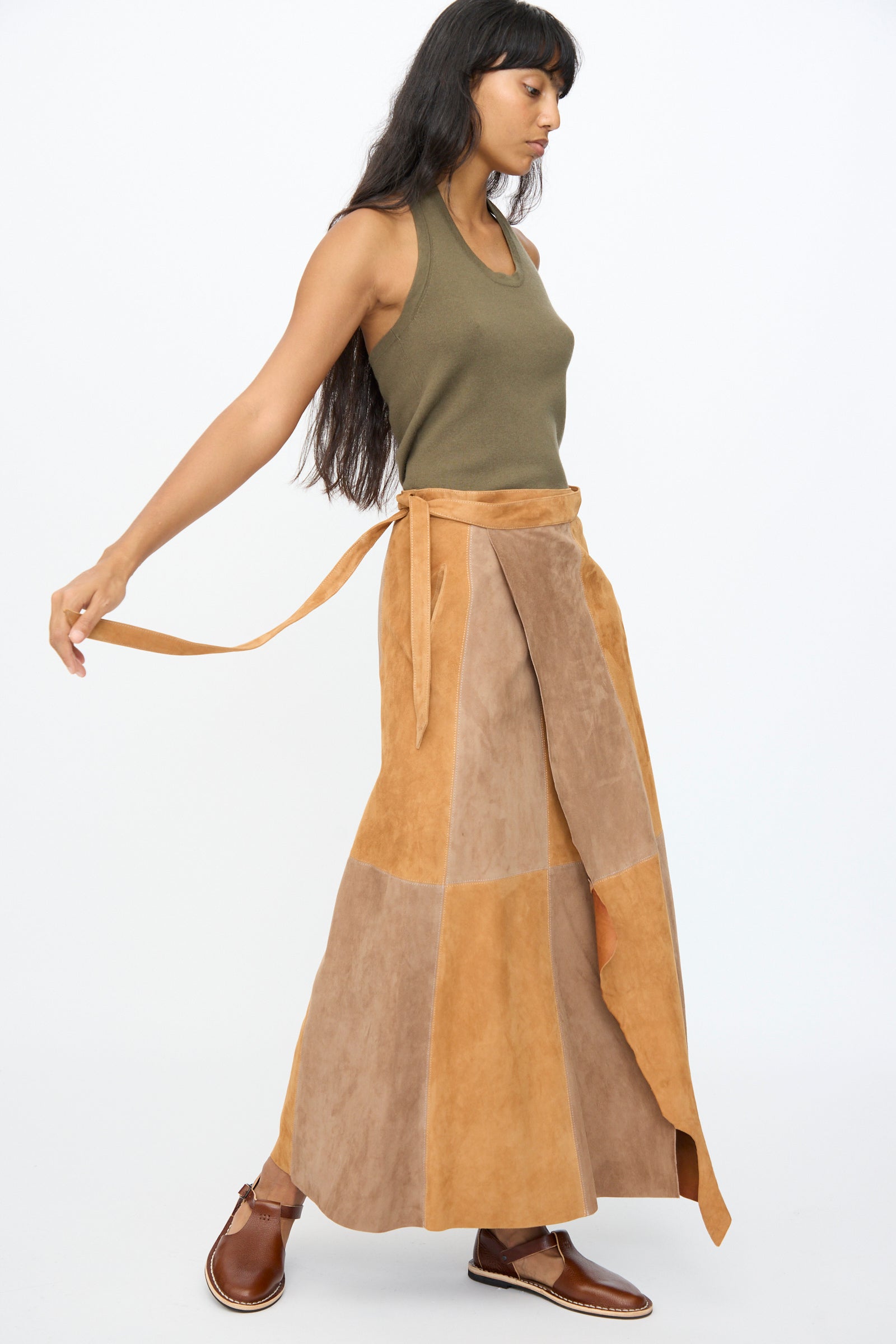A woman wearing an olive green tank top, brown shoes, and the Milena Silvano Suede Persephone Skirt in Cinnamon stands against a plain white background.