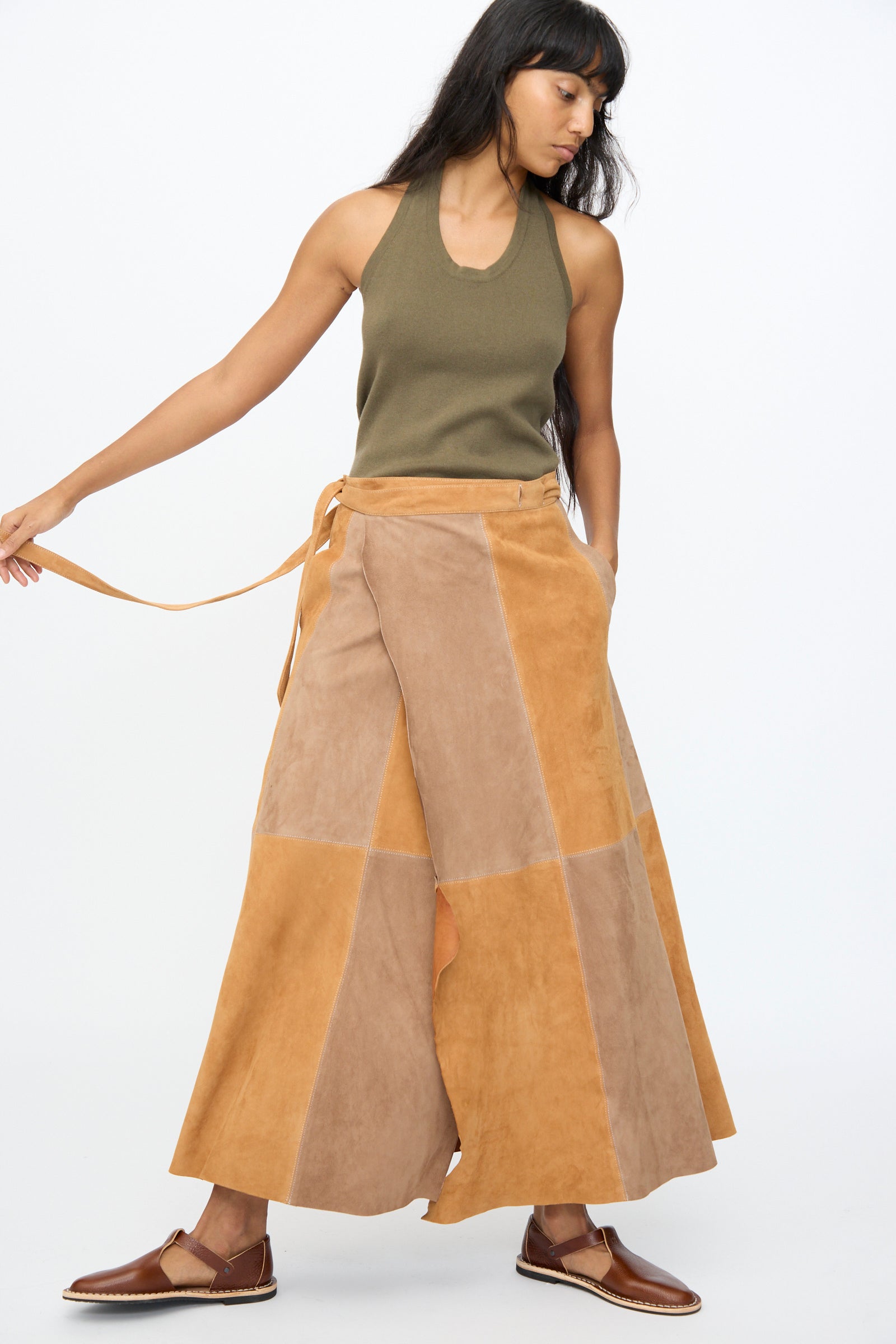 A woman wears the Milena Silvano Suede Persephone Skirt in Cinnamon, styled with a sleeveless olive top and brown flats, standing against a plain white background.