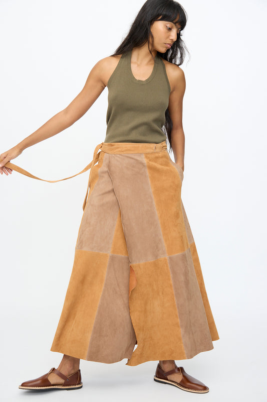 A woman wears the Milena Silvano Suede Persephone Skirt in Cinnamon, styled with a sleeveless olive top and brown flats, standing against a plain white background.