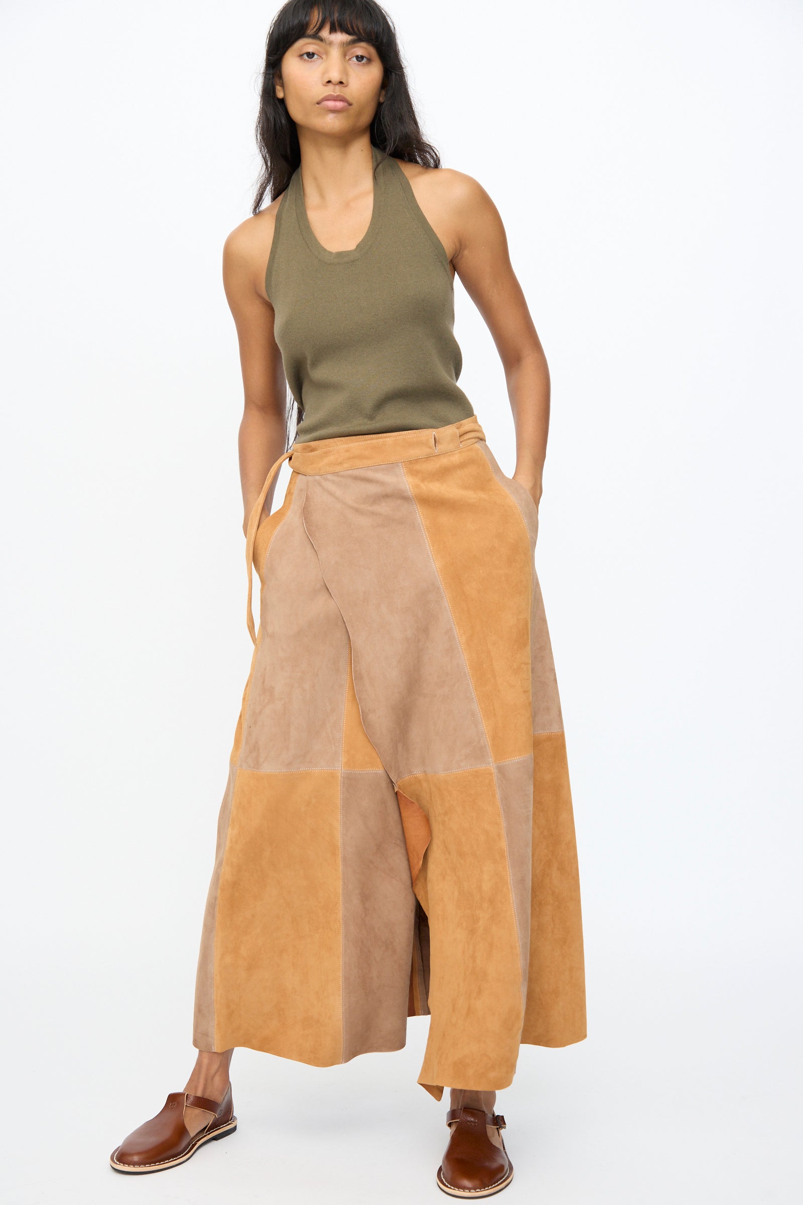 A woman stands against a plain background wearing the Suede Persephone Skirt in Cinnamon by Milena Silvano and a sleeveless olive top with brown slip-on shoes.