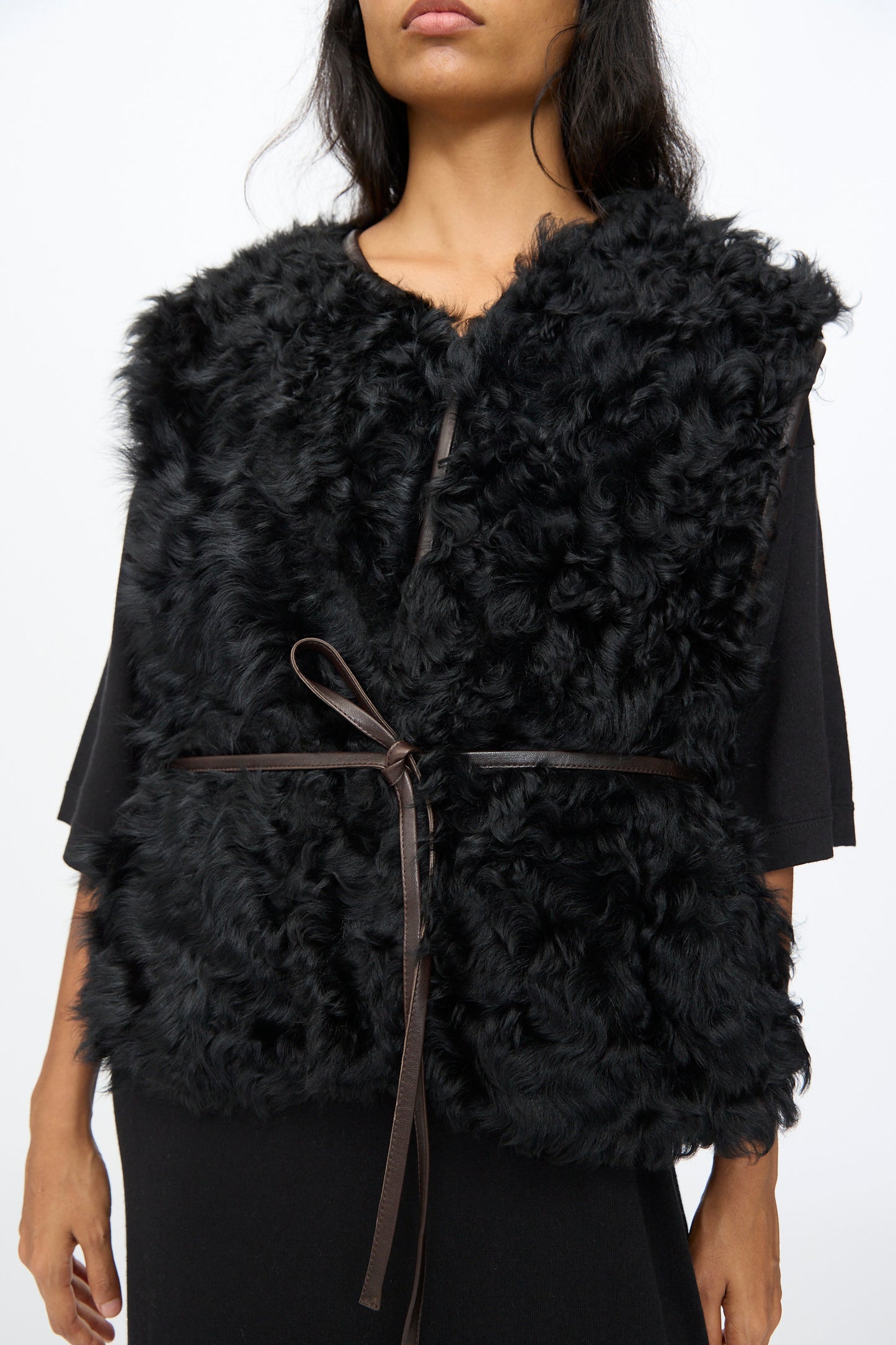 A person wearing the Milena Silvano Transition Vest in Chaga—a relaxed fit black faux fur vest with a thin waist tie—over a black top, shown from shoulders to hips.