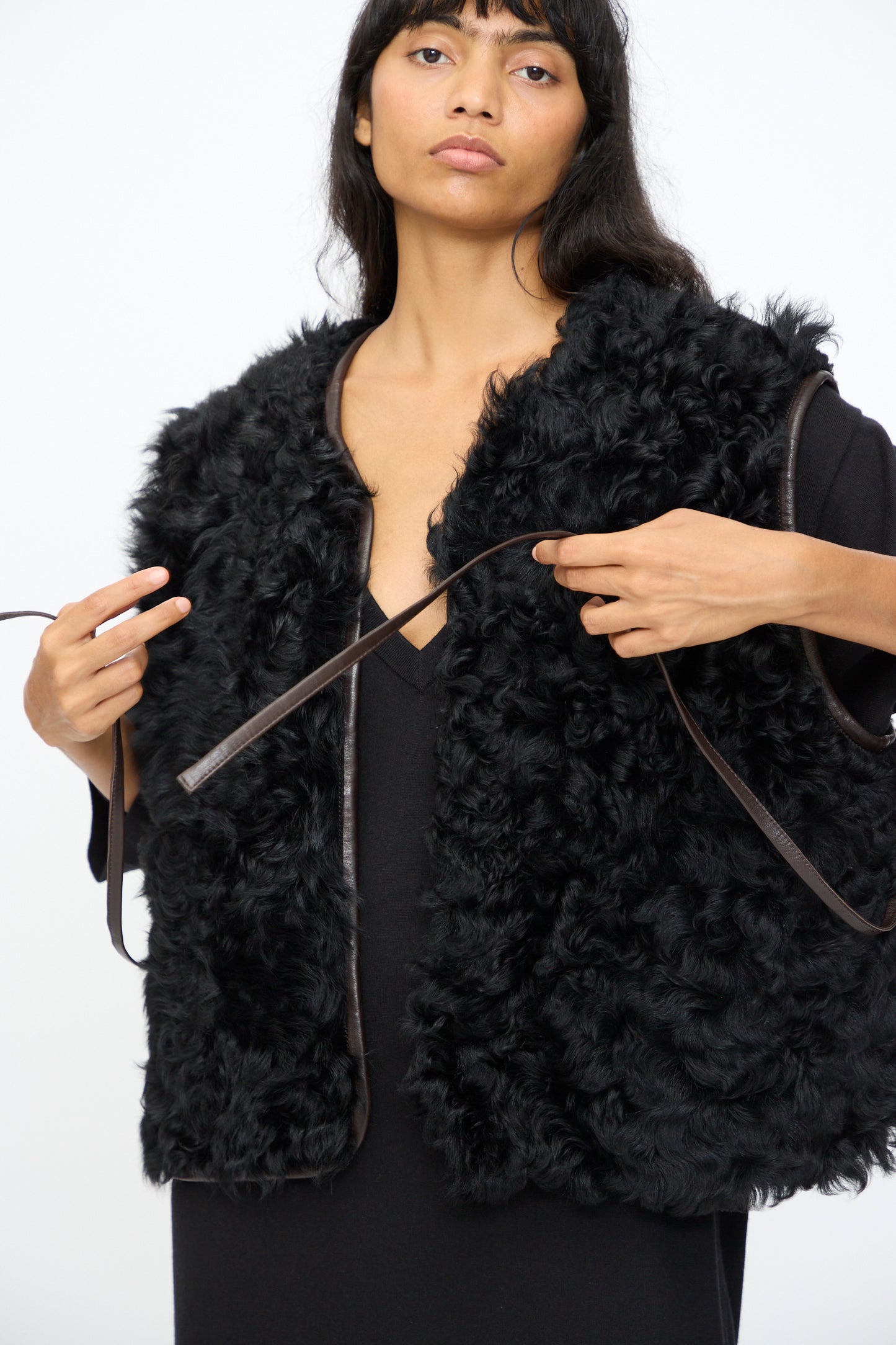 A woman wears the Milena Silvano Transition Vest in Chaga over a black top, adjusting its brown tie straps, against a plain white background.