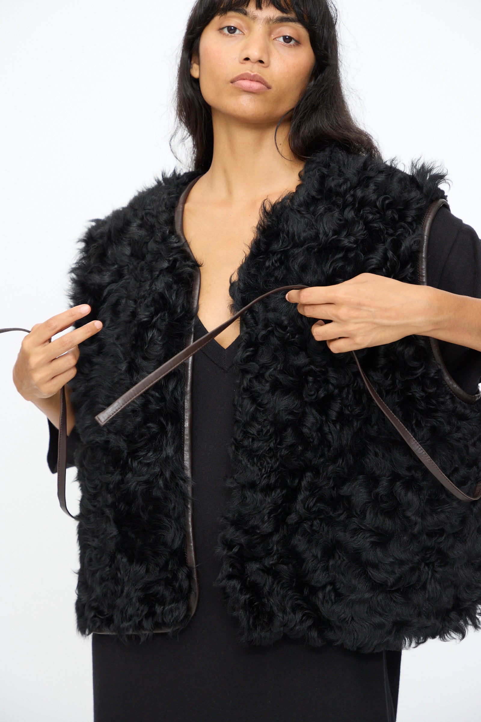 A woman wears the Milena Silvano Transition Vest in Chaga over a black top, adjusting its brown tie straps, against a plain white background.