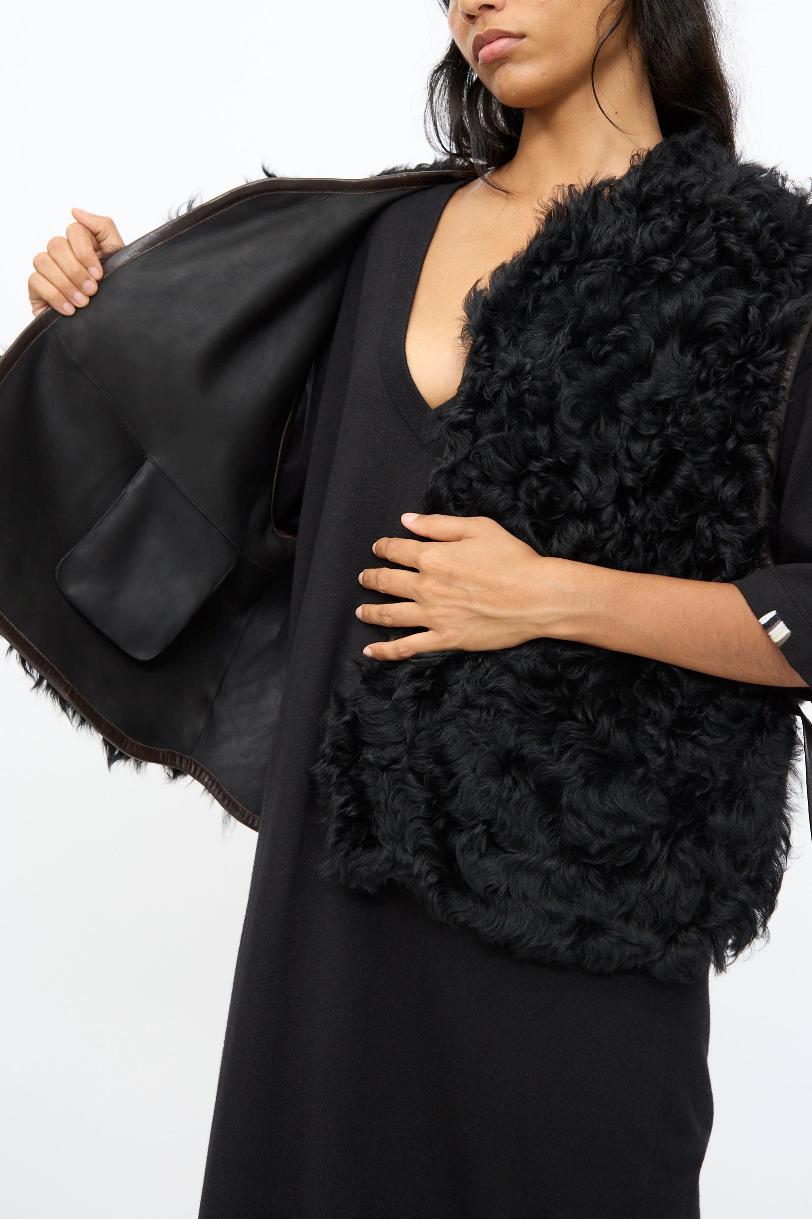 A woman wears the Milena Silvano Transition Vest in Chaga—textured black sheepskin with a relaxed fit, open on one side to show the inner lining and pocket, crafted in Turkey, styled over a black dress.