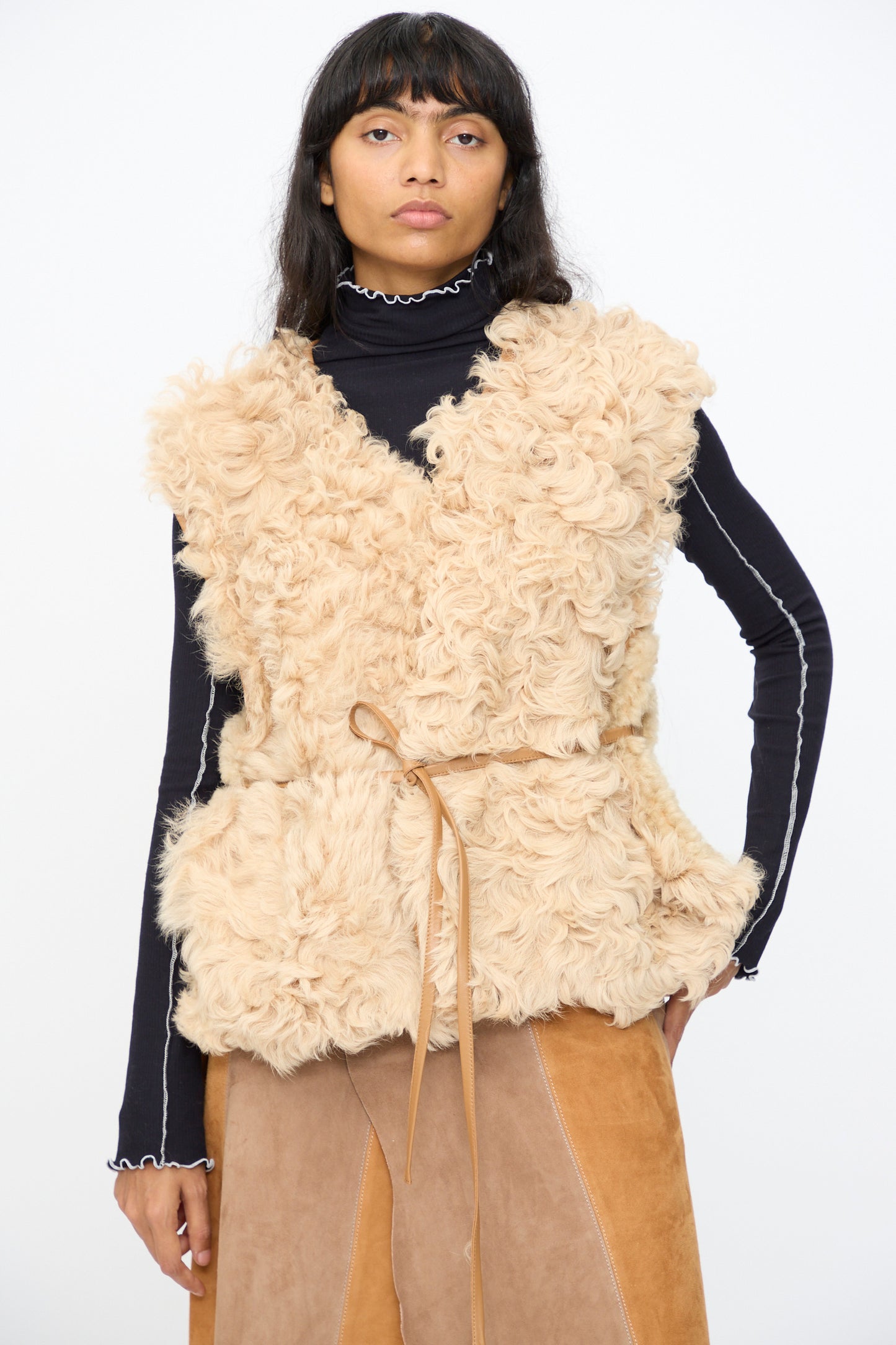 A person wears the Milena Silvano Transition Vest in Oat over a black turtleneck with white trim and a brown skirt, standing against a plain white background.