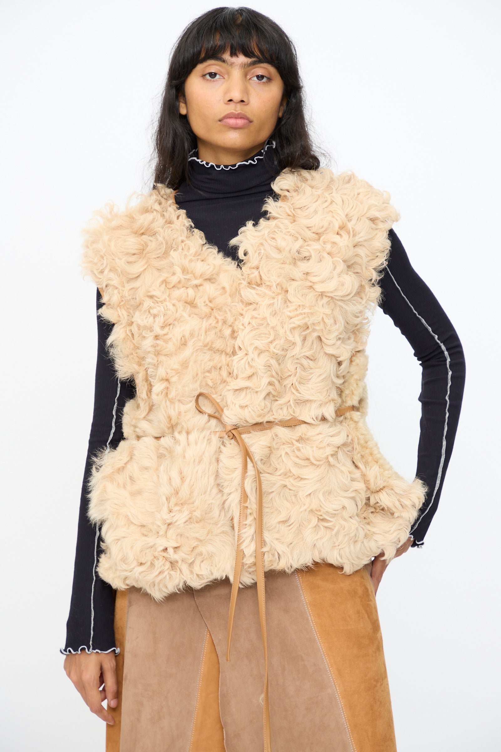 A person wears the Milena Silvano Transition Vest in Oat over a black turtleneck with white trim and a brown skirt, standing against a plain white background.