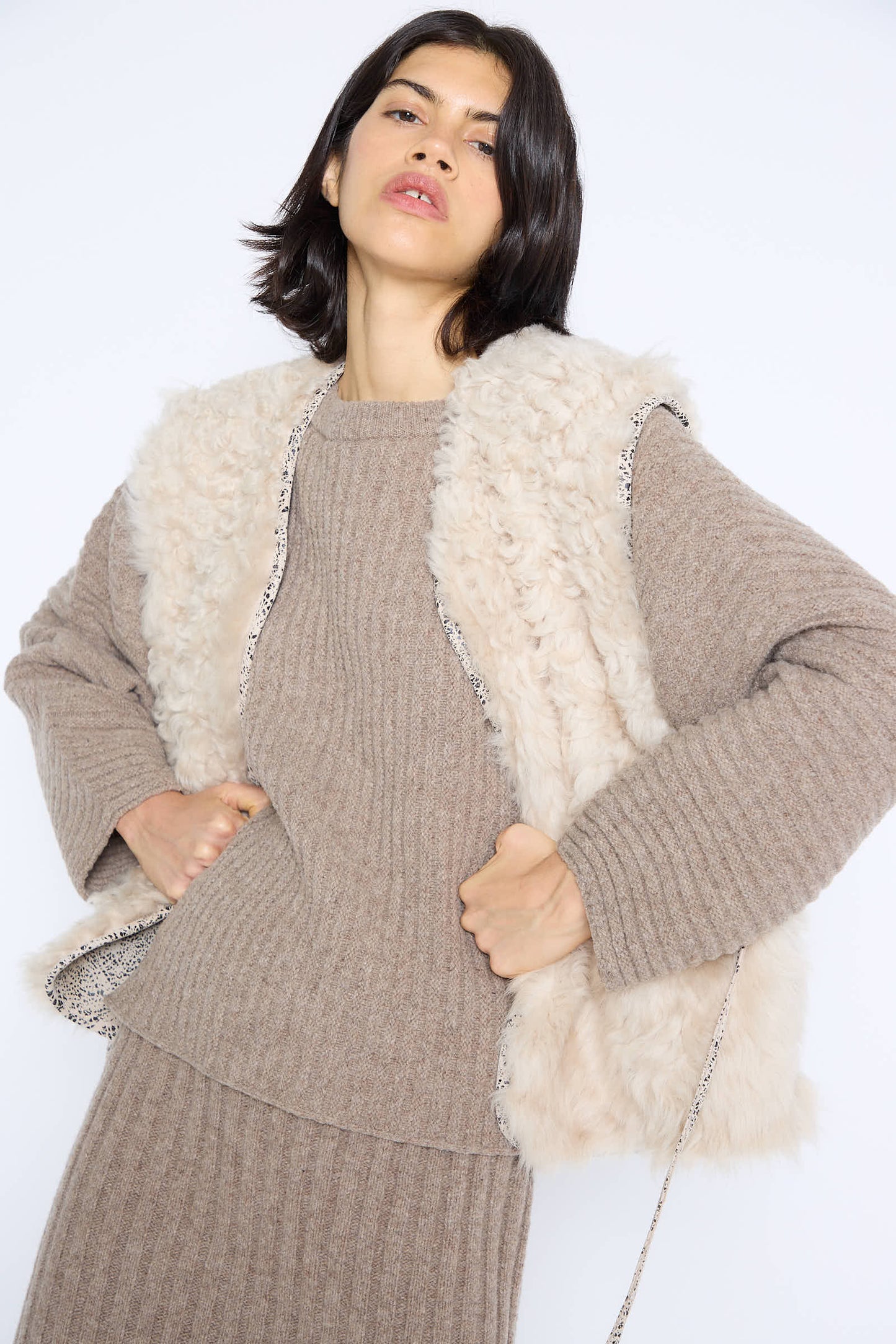 A person with dark hair wears a beige knit sweater and matching pants paired with Milena Silvano's Transition Vest in Classic, standing with hands on hips against a plain background.