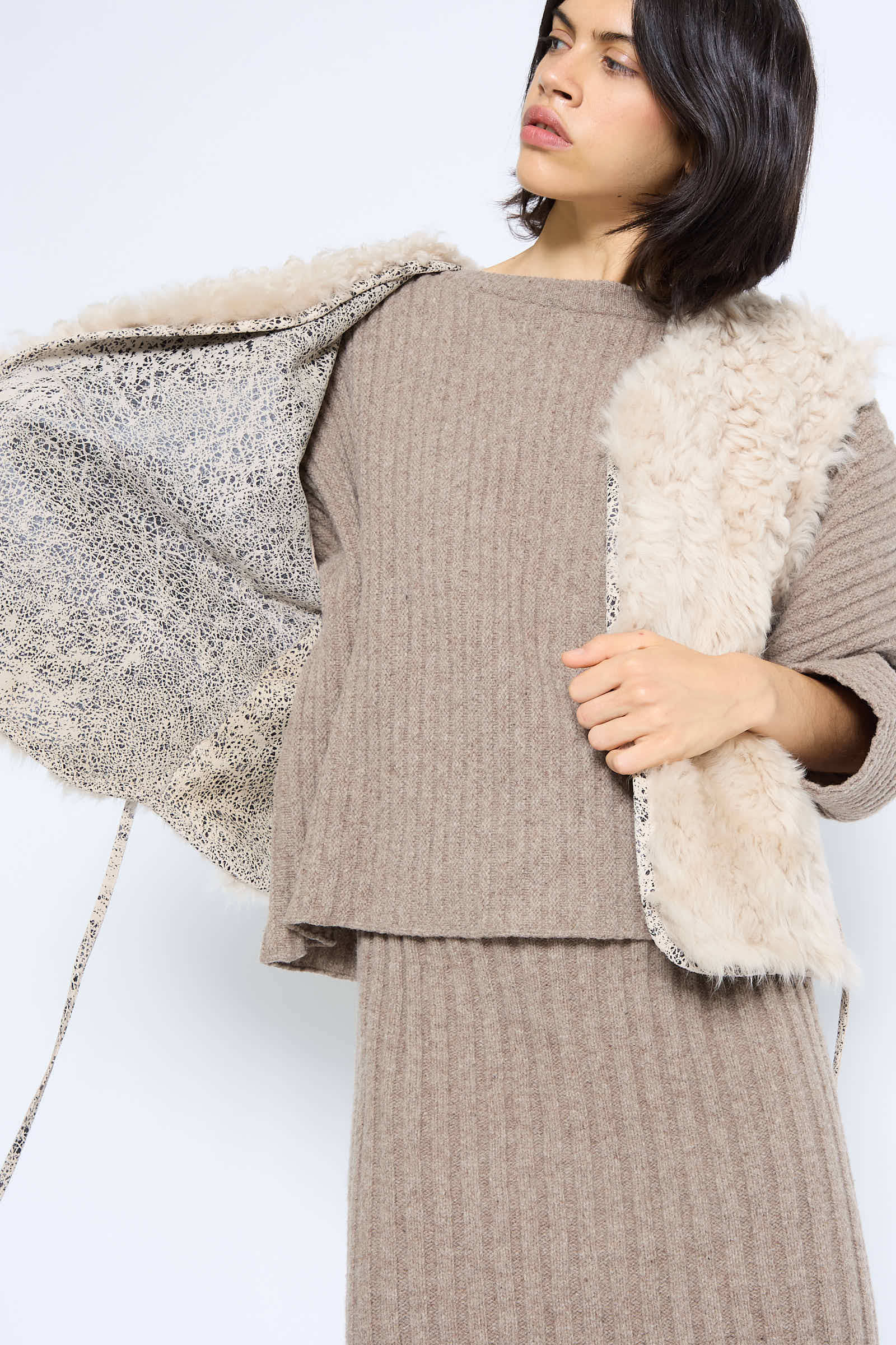 A woman wearing a beige knitted outfit adjusts the Milena Silvano Transition Vest in Classic, featuring tonal textures and a patterned inner lining.